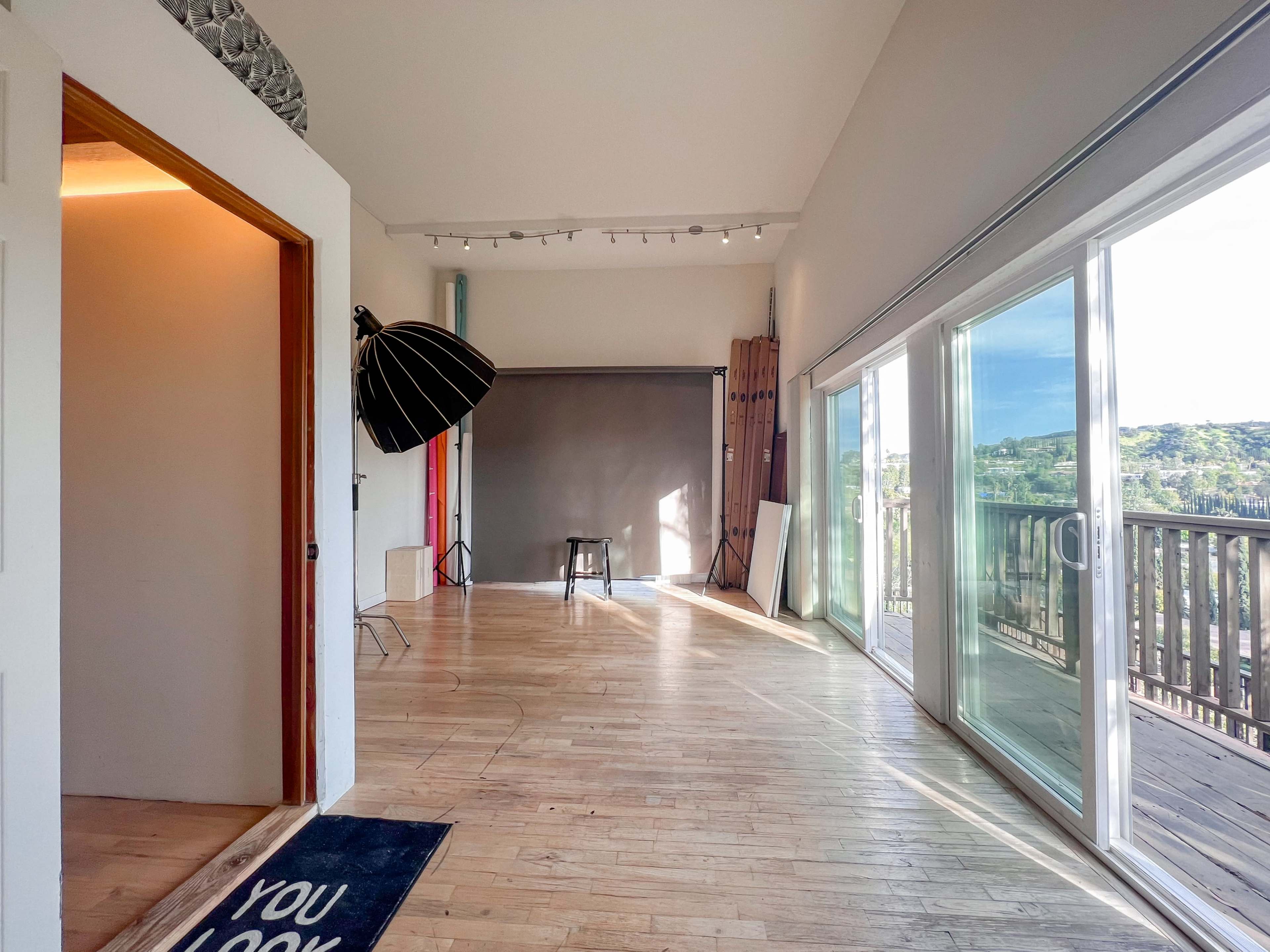 The image shows a spacious, well-lit room with large windows, a wooden floor, and photography equipment, including a large umbrella light and a stool.