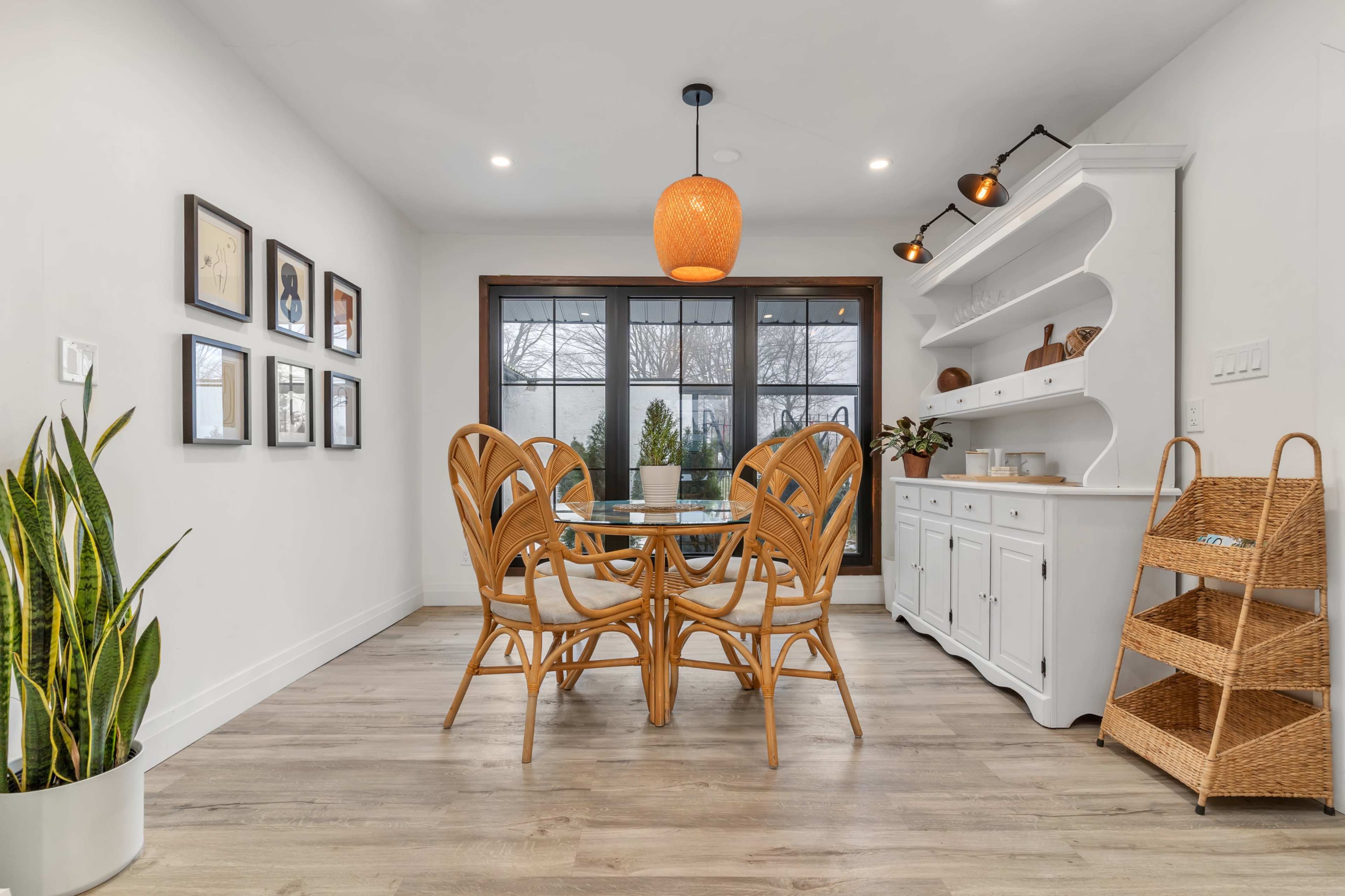 This image shows a dining area featuring a round glass table surrounded by four rattan chairs, with a white sideboard and shelves against the wall, and a large window allowing natural light to enter.