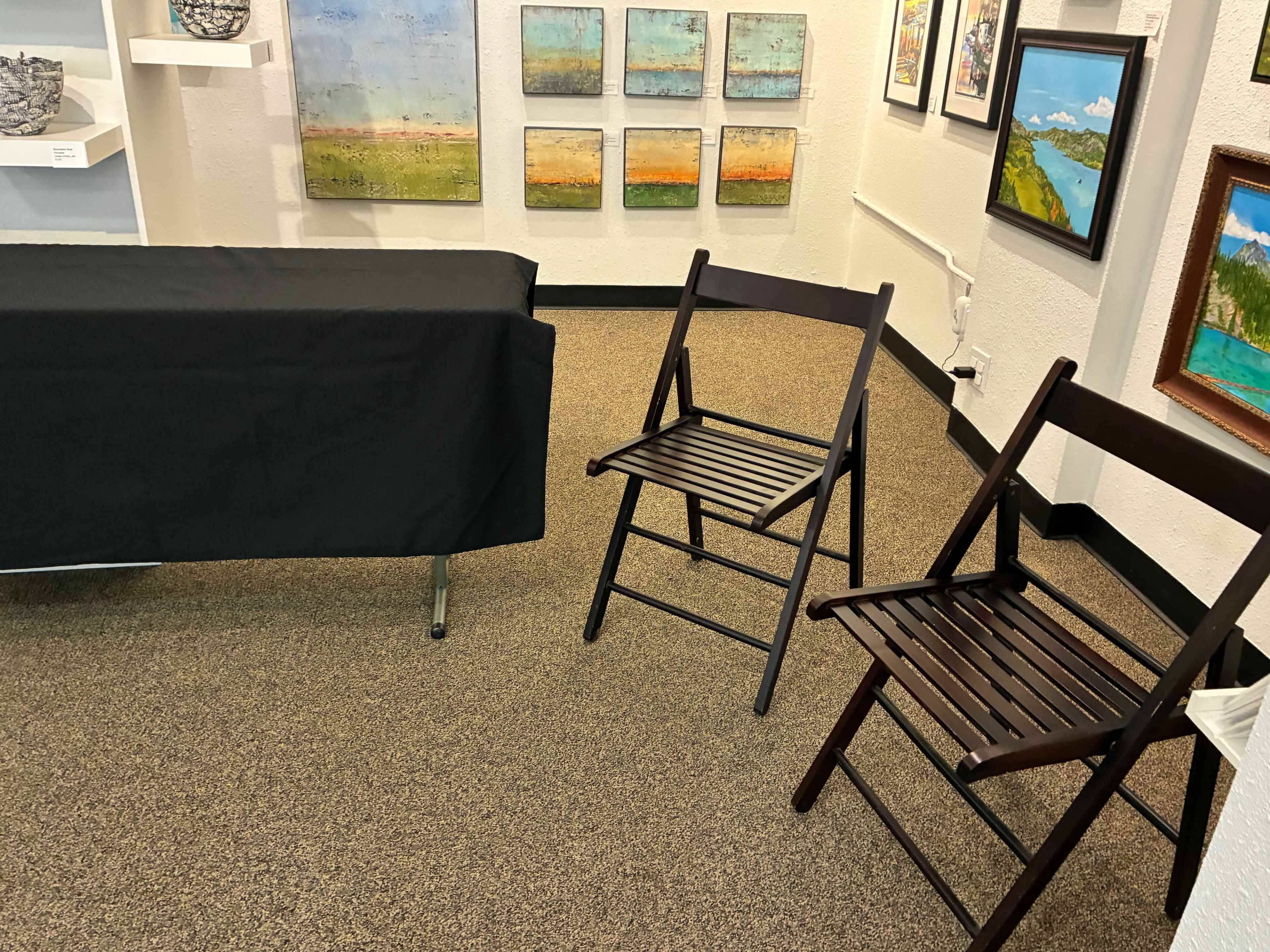 The image shows a small gallery or exhibition space with a table covered by a black cloth and two wooden folding chairs on carpeted flooring, surrounded by various framed artworks on the walls.