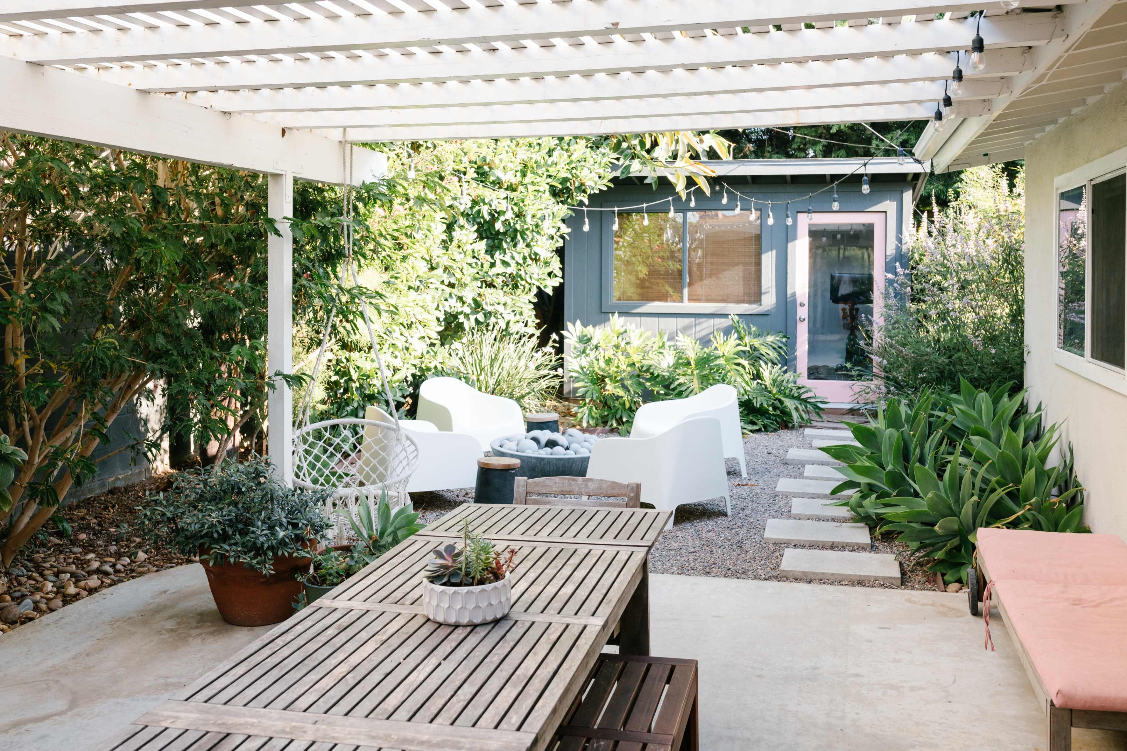 A patio area features a wooden table, white chairs, and a pathway leading to a small garden shed, surrounded by lush greenery.