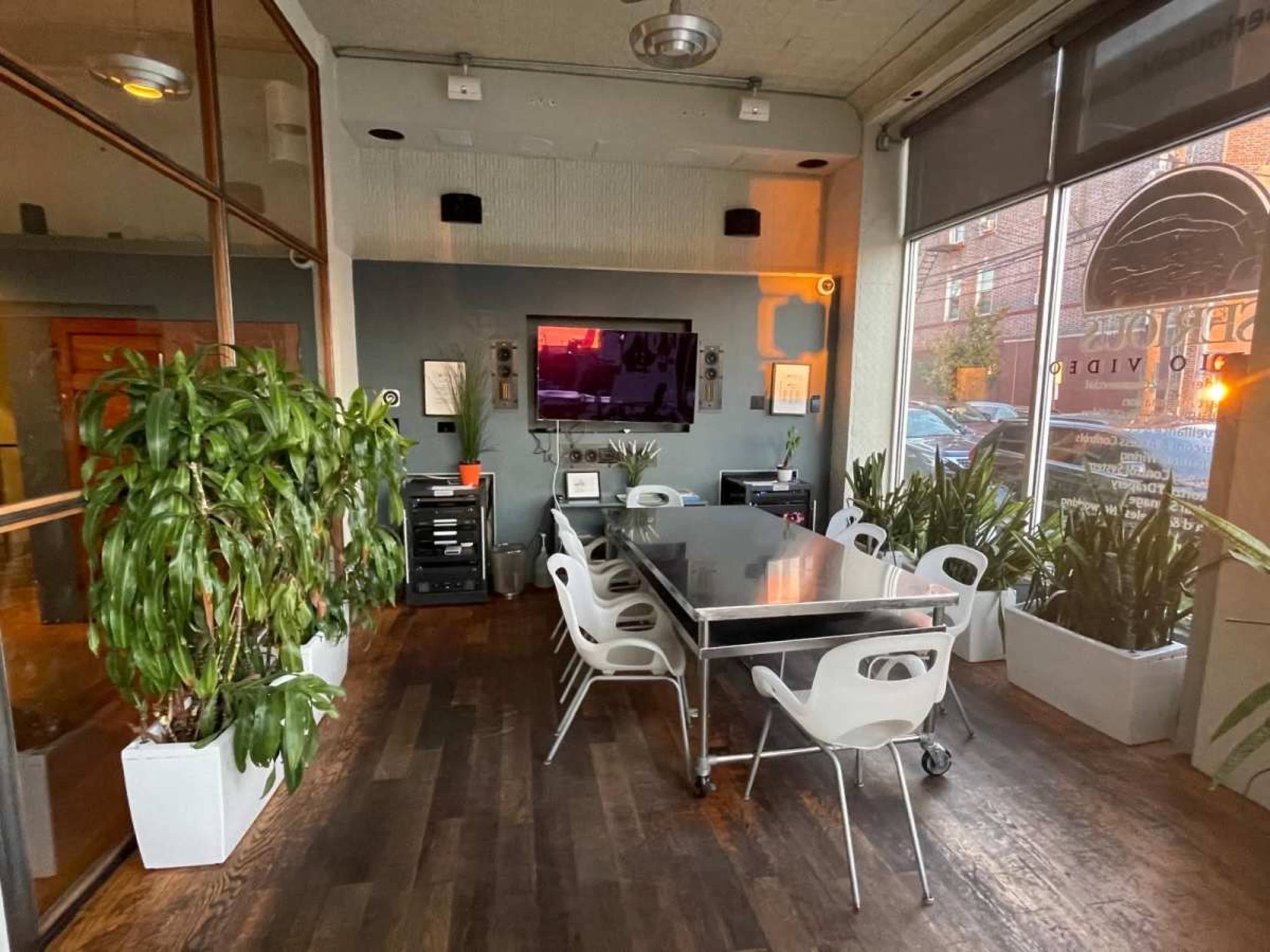 A dining area features a metal table surrounded by white chairs, with large potted plants and a television mounted on the wall.