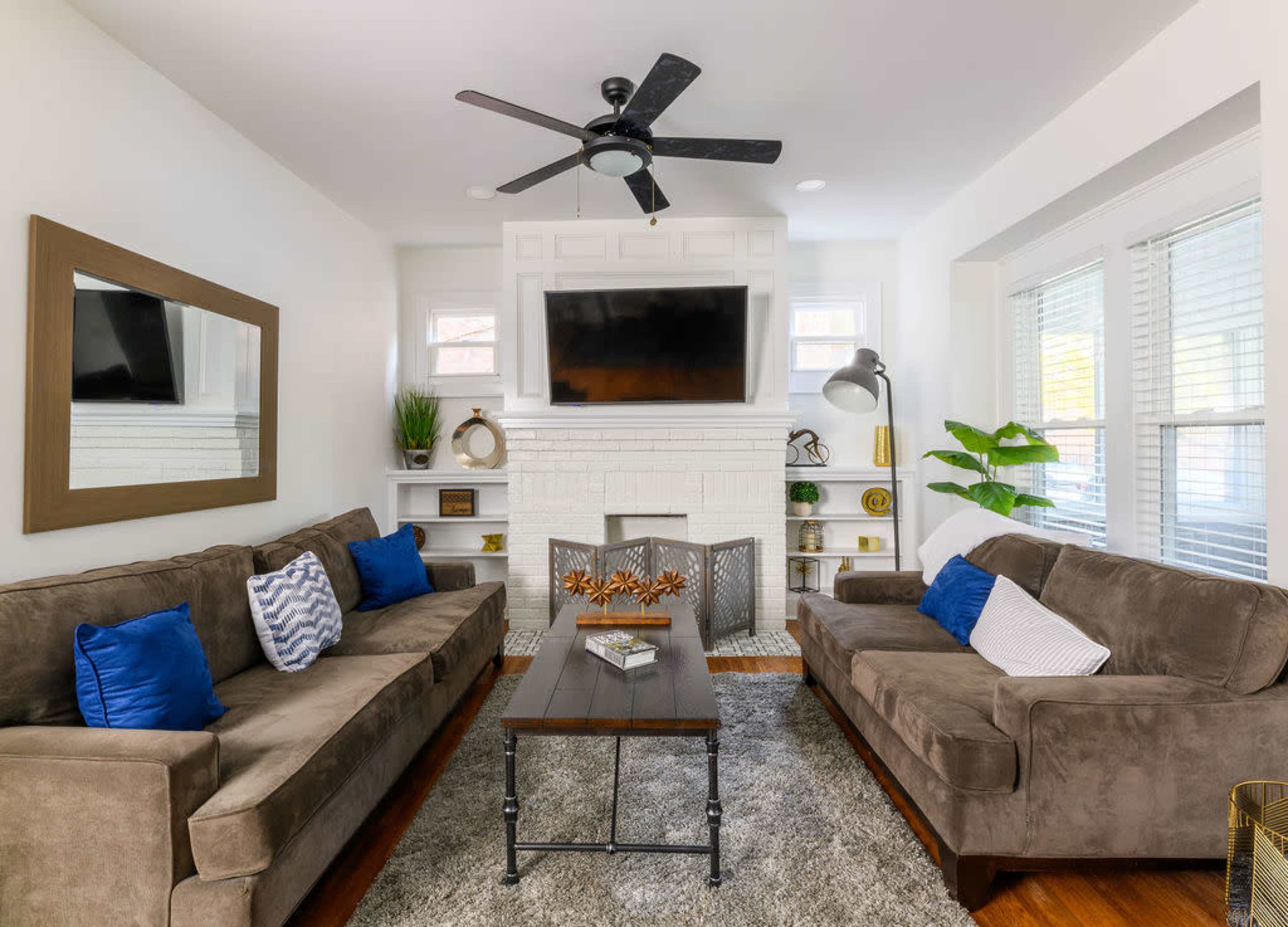A living room features two brown sofas with blue and white pillows, a coffee table in the center, and a television mounted above a white brick fireplace, with large windows allowing natural light.