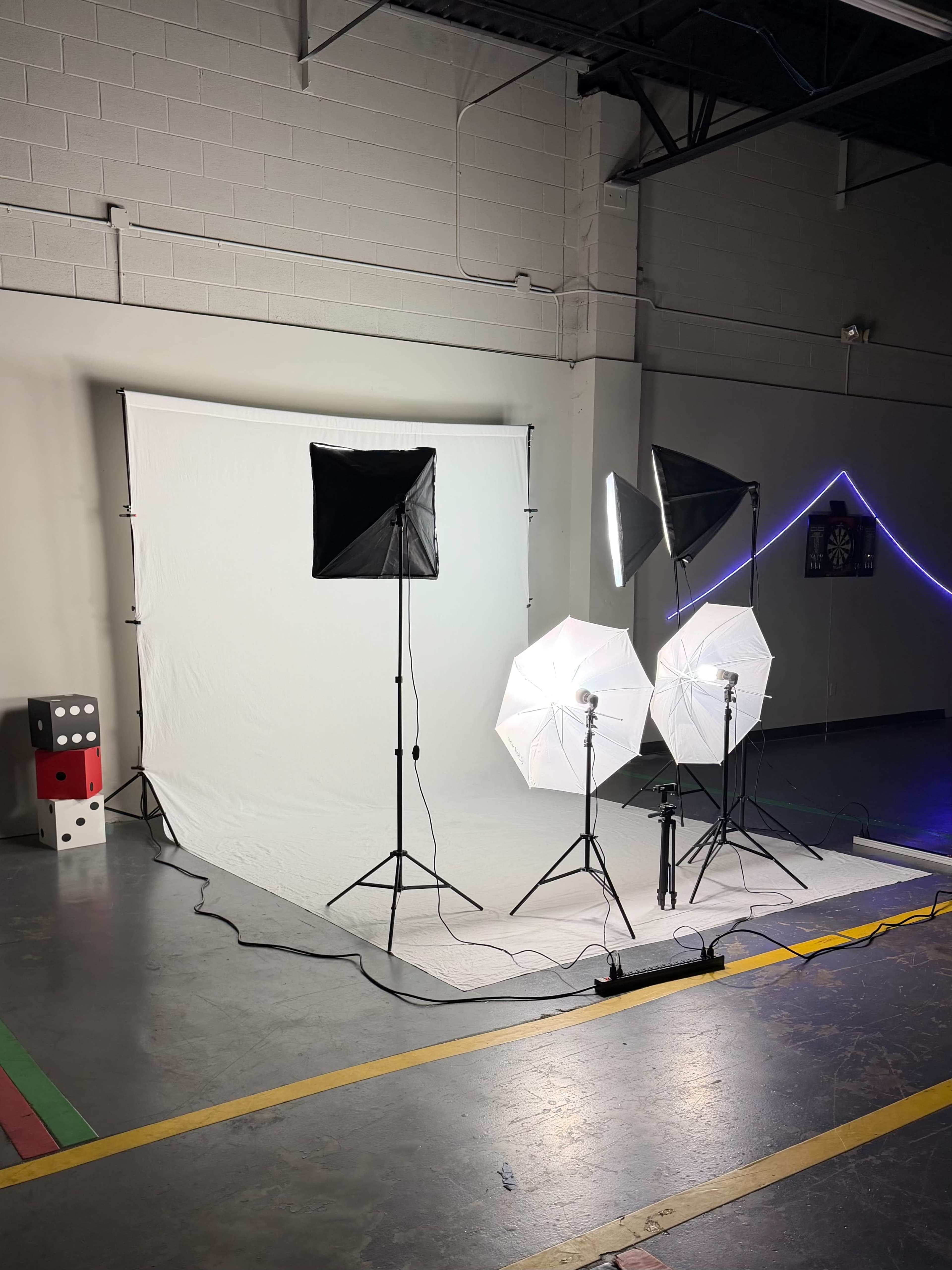 The image shows a photography studio setup with a white backdrop, three lighting equipment units, and props in the background.