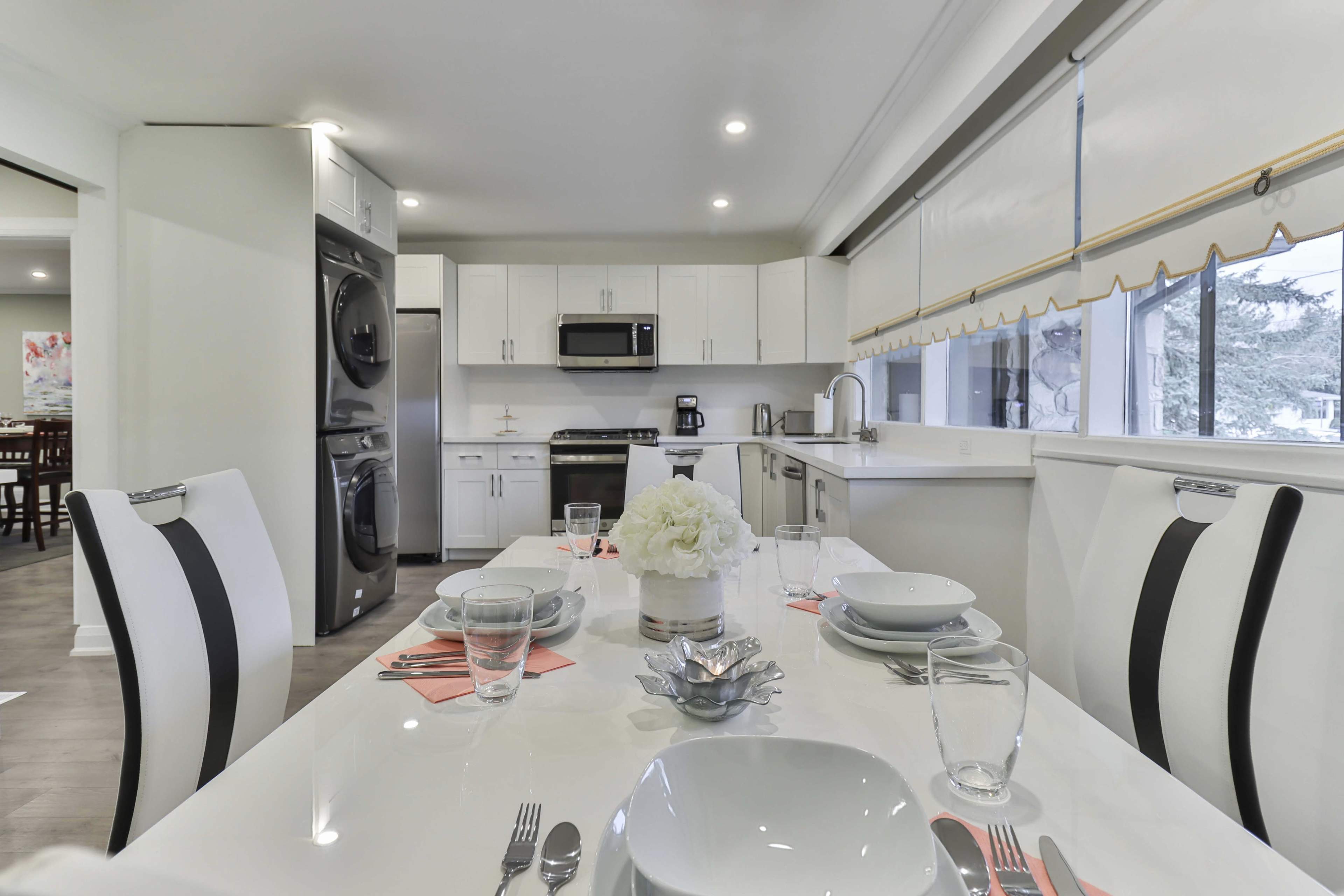 A modern kitchen features a dining table set for a meal, with white dishes and glassware, alongside appliances and cabinets in a sleek design.