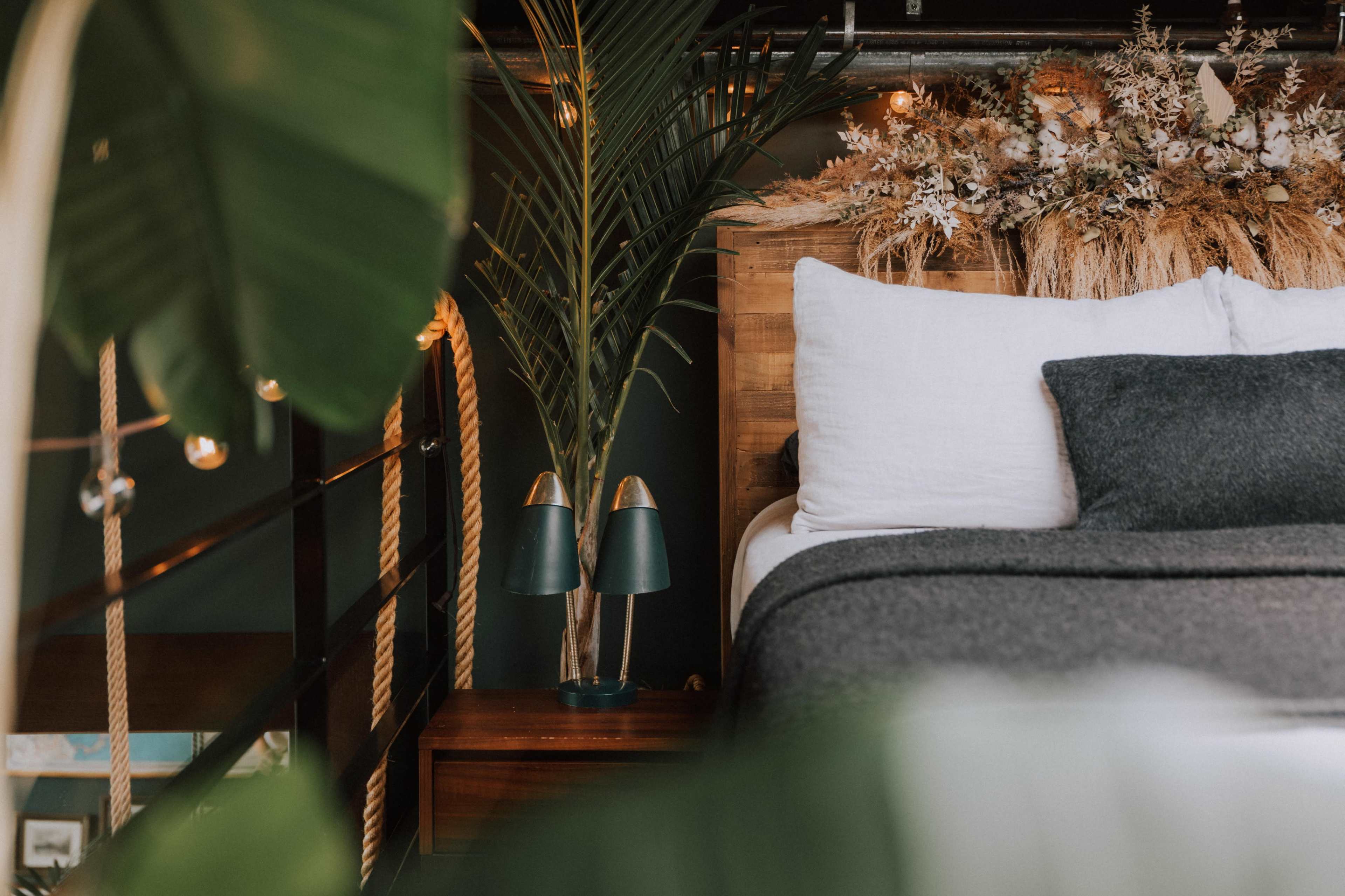 A cozy bedroom features a bed with white pillows and a gray blanket, accompanied by a bedside table with green lamps and decorative plants.