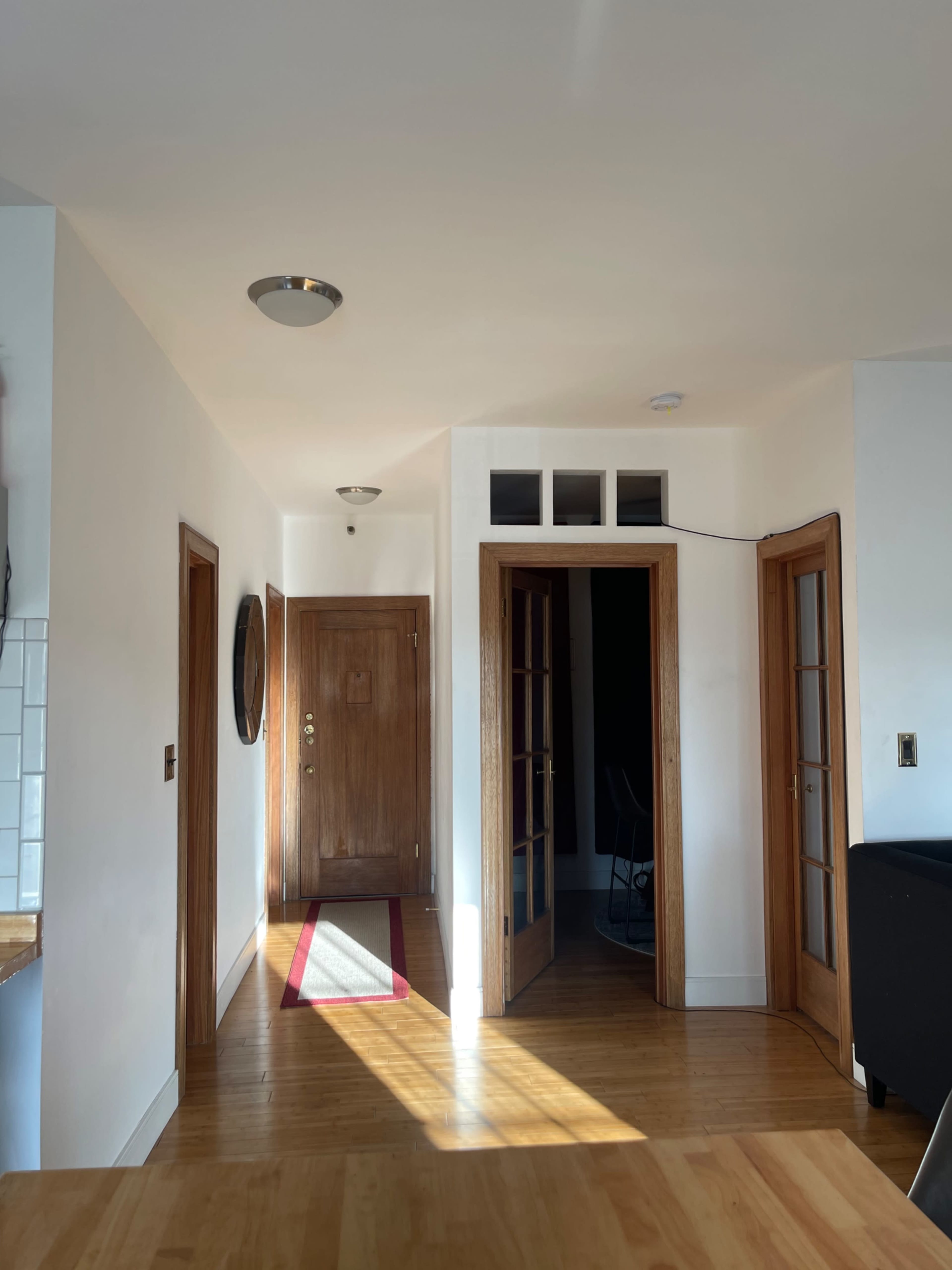 A bright interior hallway with wooden doors, a light fixture, and a striped rug leading to an entrance door.