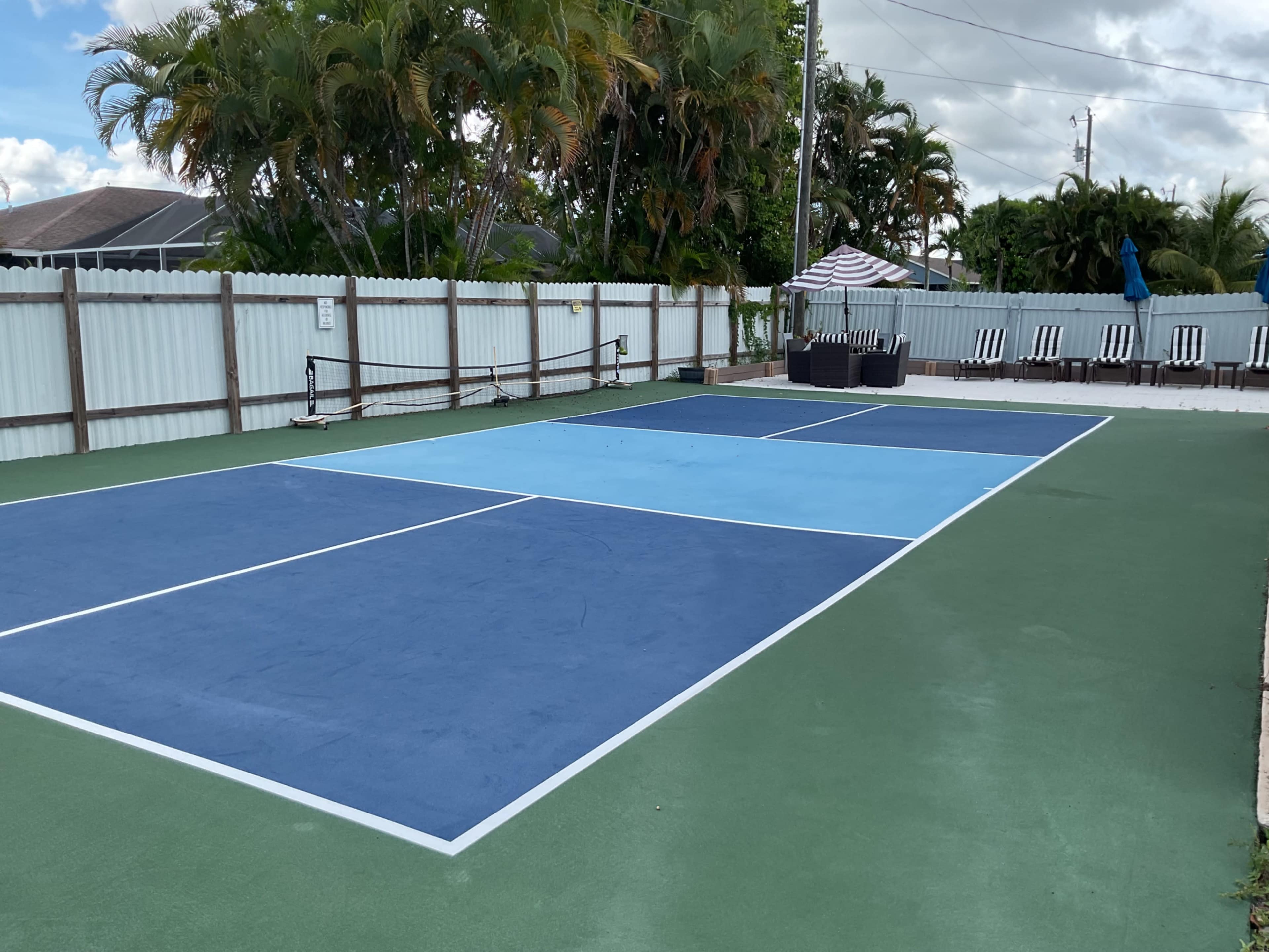 The image shows a tennis court with blue and green playing surfaces surrounded by a white fence and tropical plants.