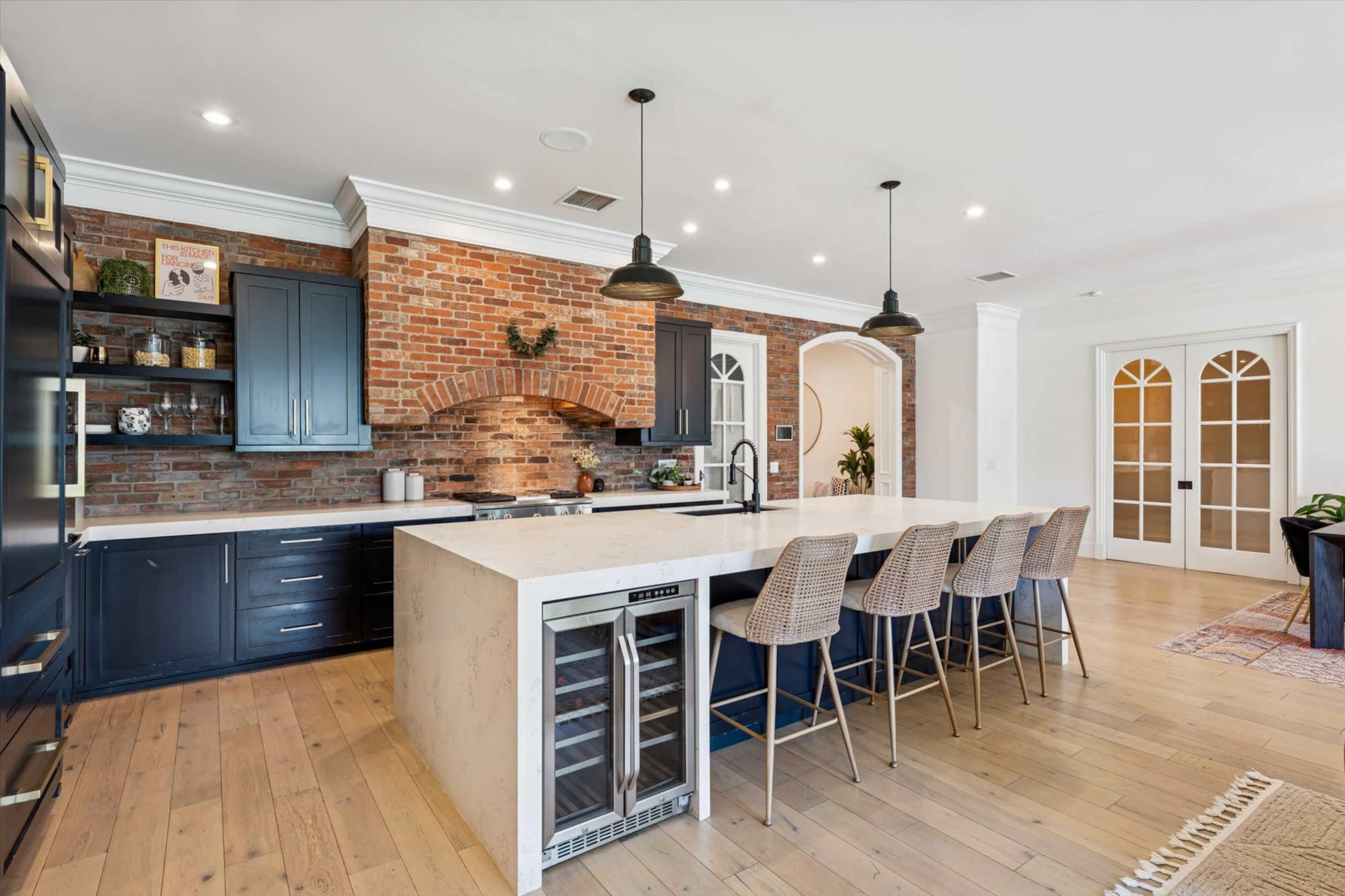 The image shows a modern kitchen featuring a large island with seating, a brick backsplash, dark cabinetry, and wooden flooring.