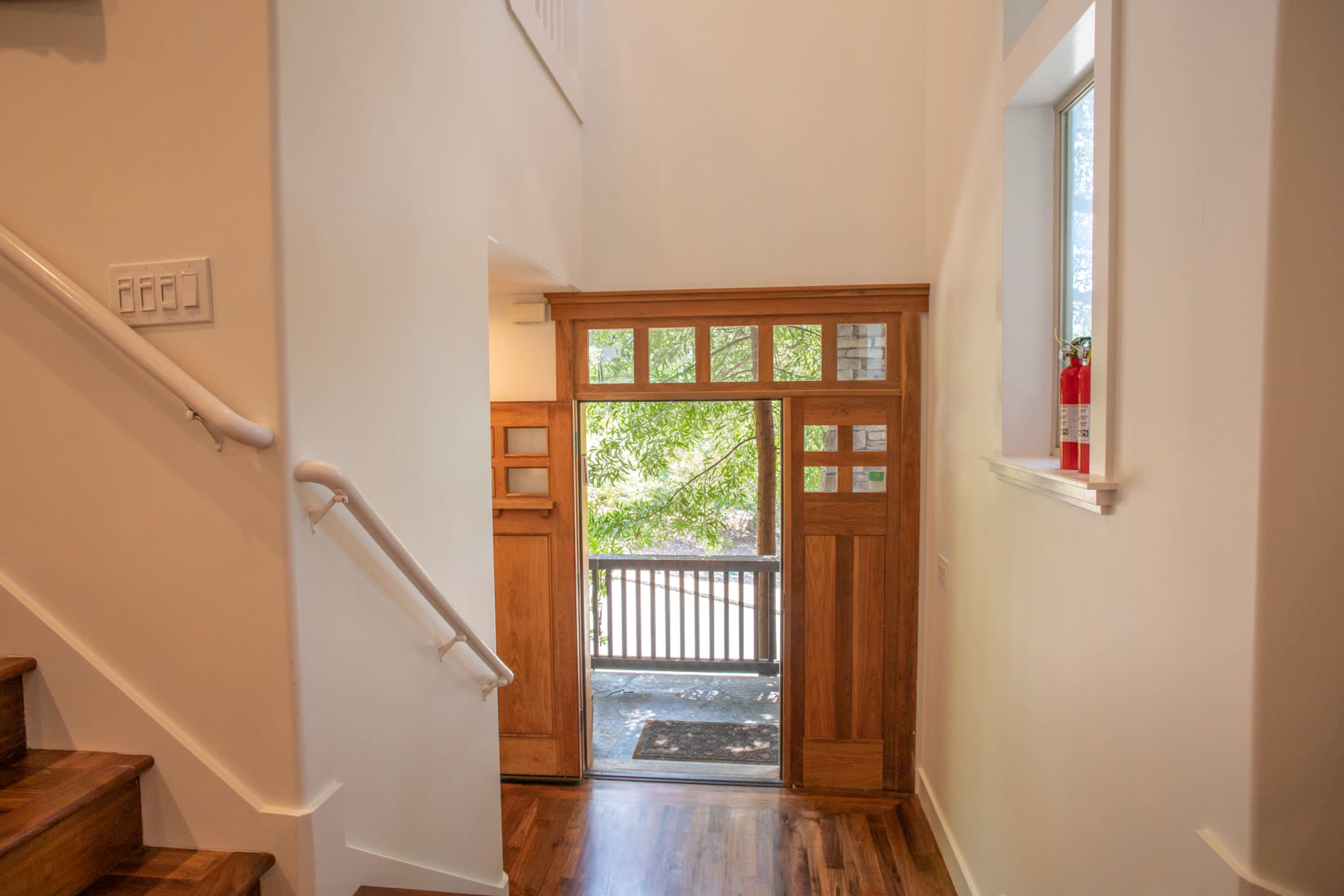 The image shows a modern entryway with wooden stairs leading up and a large doorway framed by glass panes, allowing light to enter from outside.