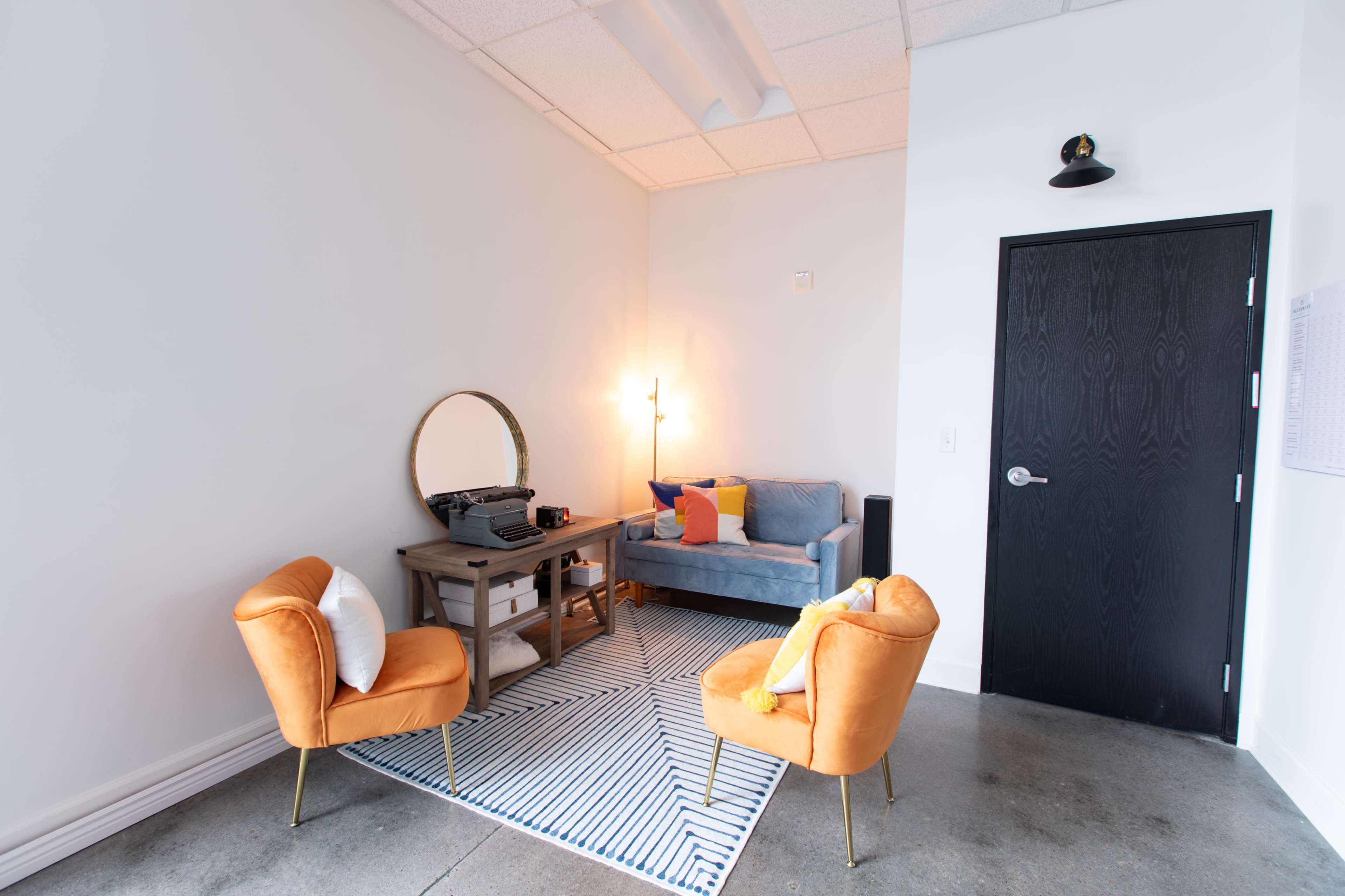 A small, modern lounge area with two orange chairs, a gray sofa, a round mirror, and a vintage typewriter on a table, set against white walls and a concrete floor.