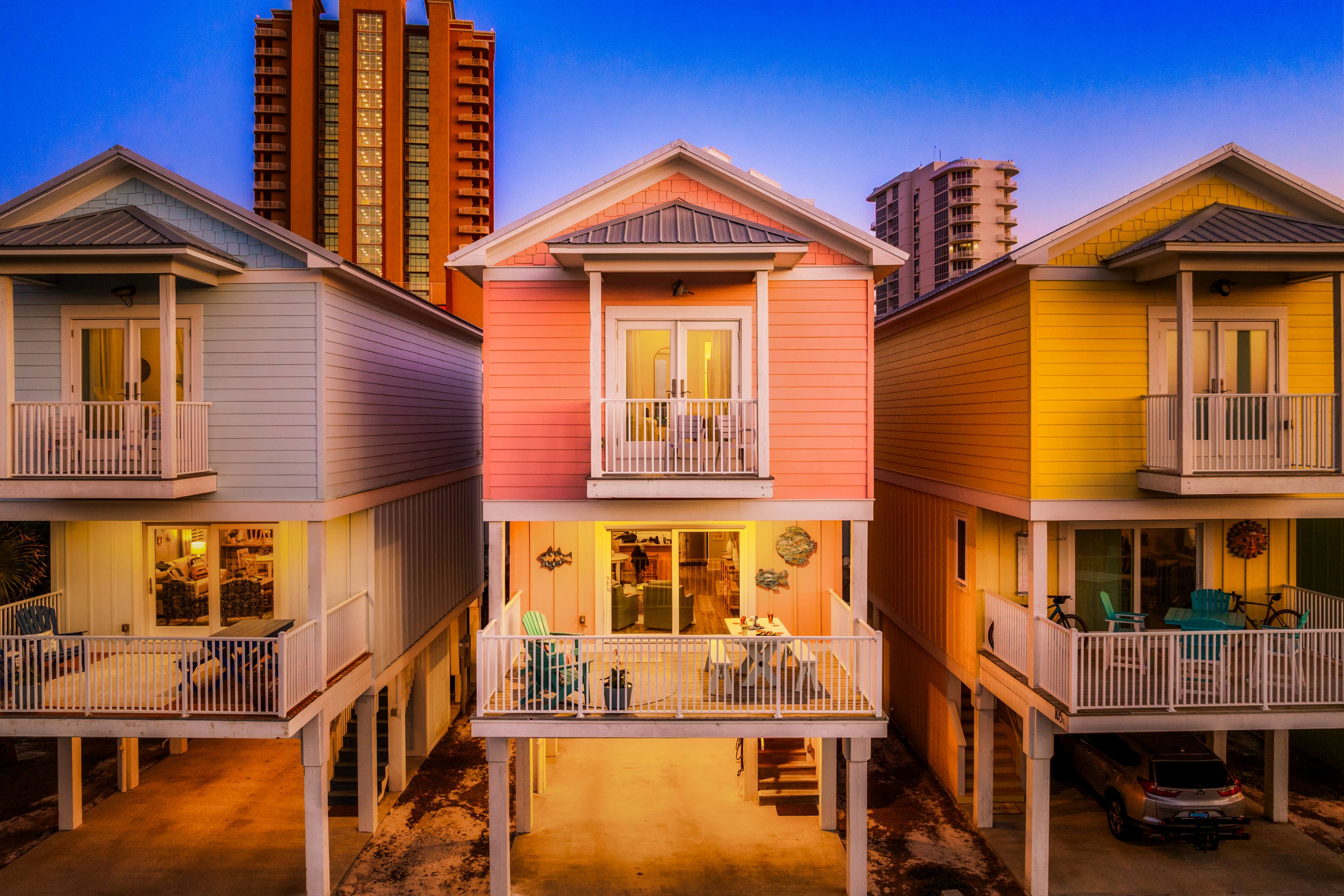 The image shows a row of colorful beachfront houses, with a gradient of pastel hues, set against a twilight sky and high-rise buildings in the background.