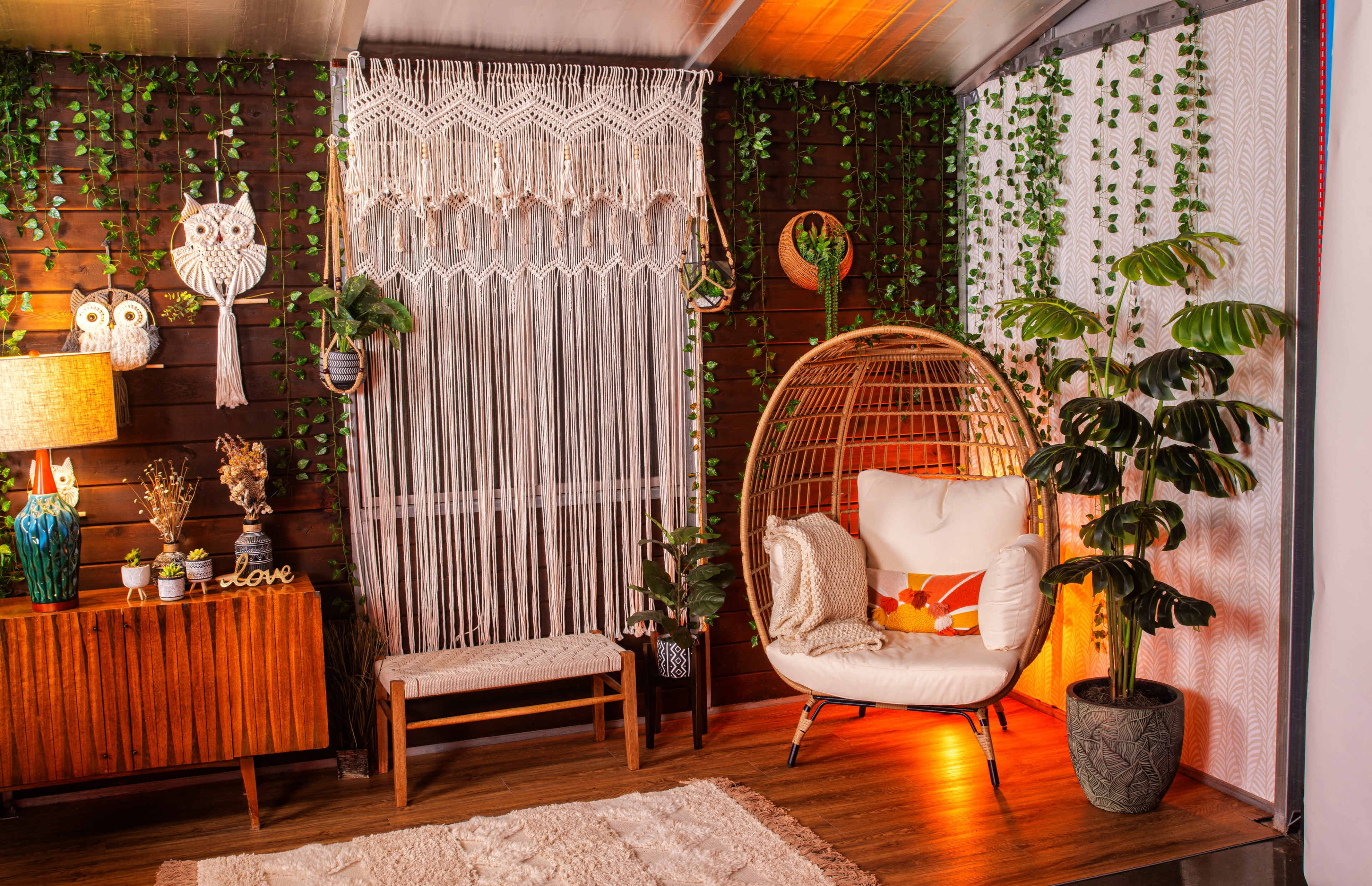 The image shows a cozy interior space featuring a rattan hanging chair, a wooden cabinet with decorative items, and lush green plants against a backdrop of macramé wall hangings and curtains.