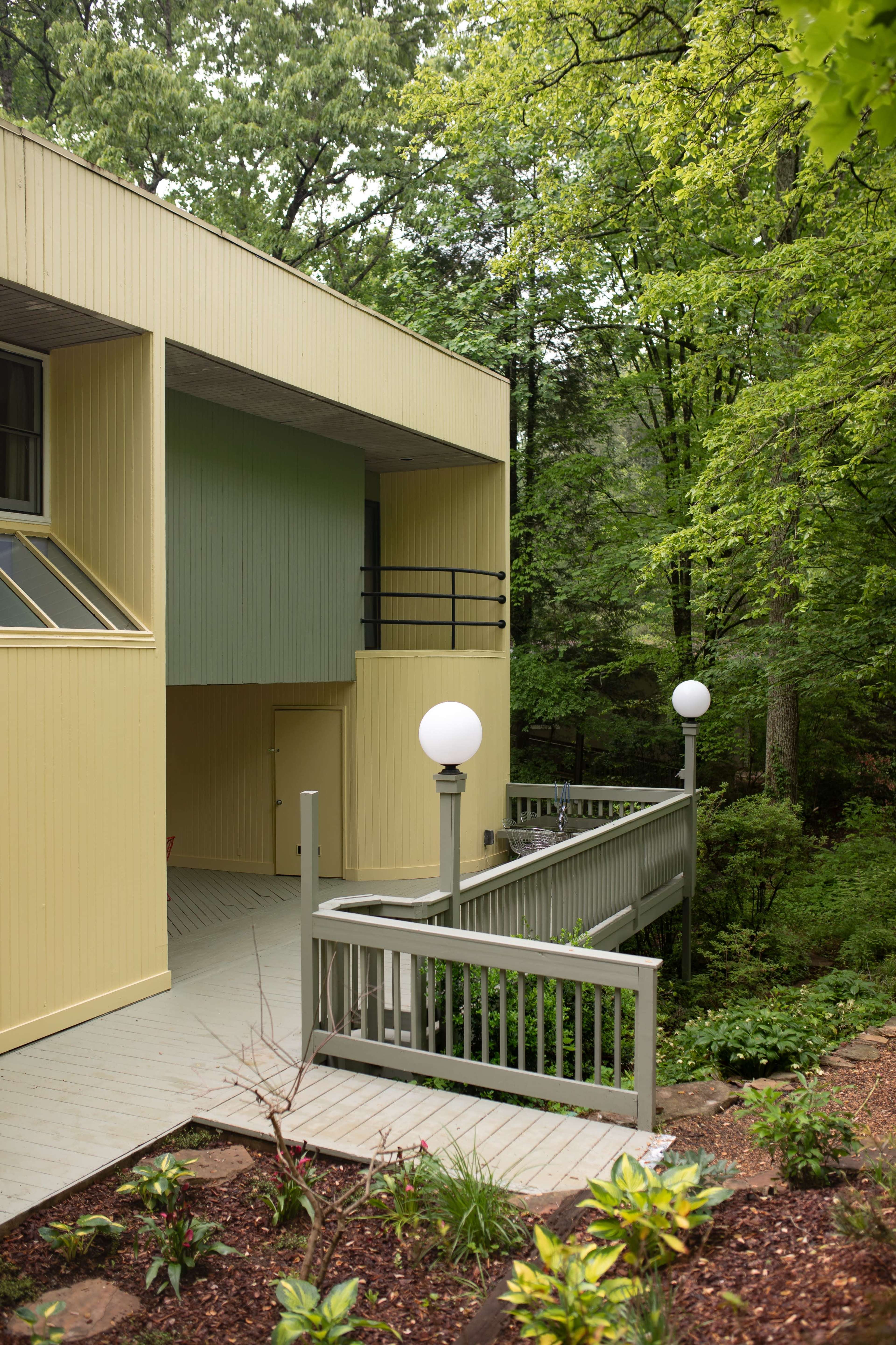 A modern building with a yellow exterior is set amidst lush green trees, featuring a pathway with spherical lamps leading to its entrance.