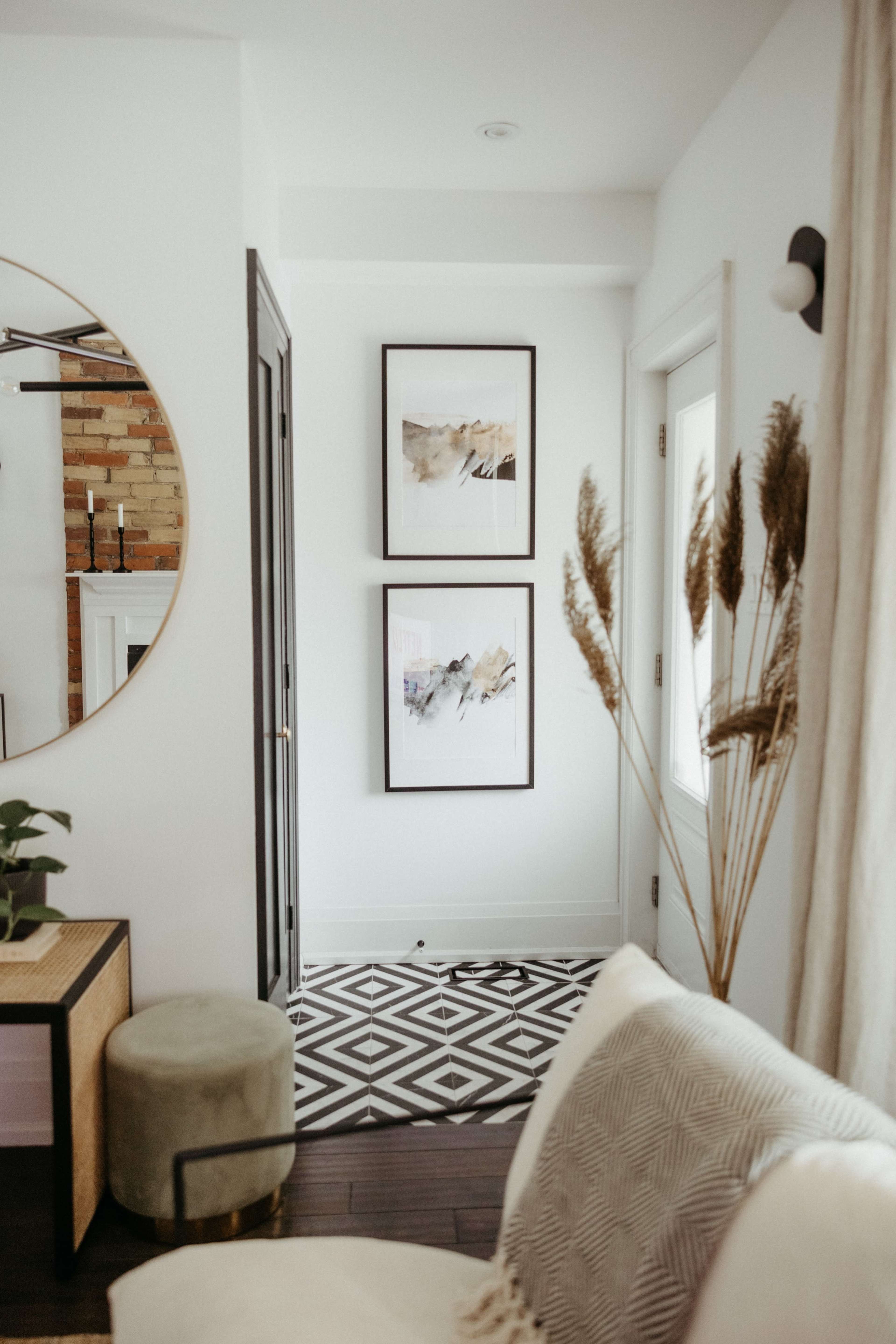 A modern interior with a corridor featuring two framed artworks on the wall, a round mirror, and decorative elements like a plant and a pouf.