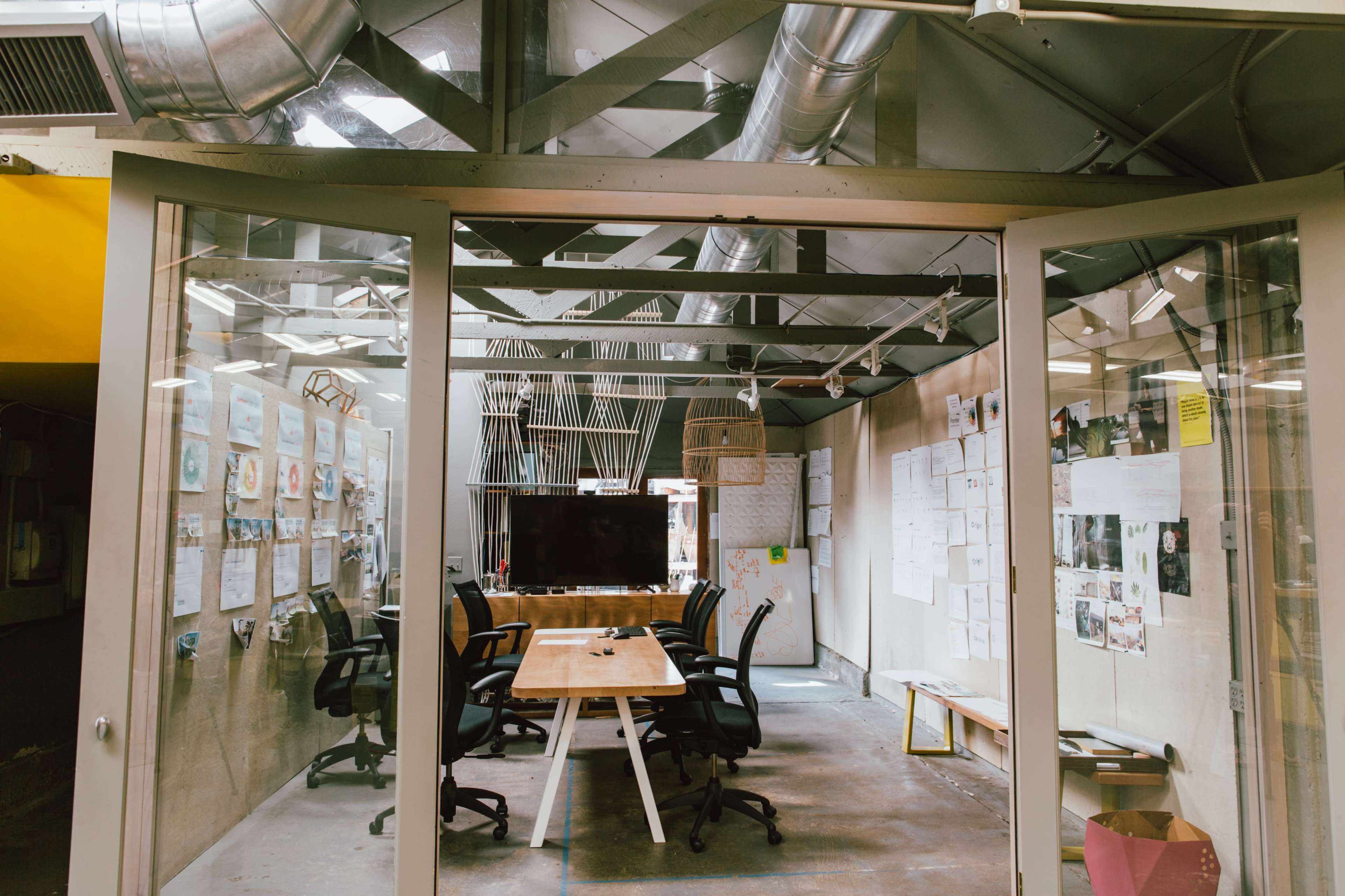 A meeting room with a wooden table and chairs is visible through large glass panels, surrounded by walls covered in papers and plans.