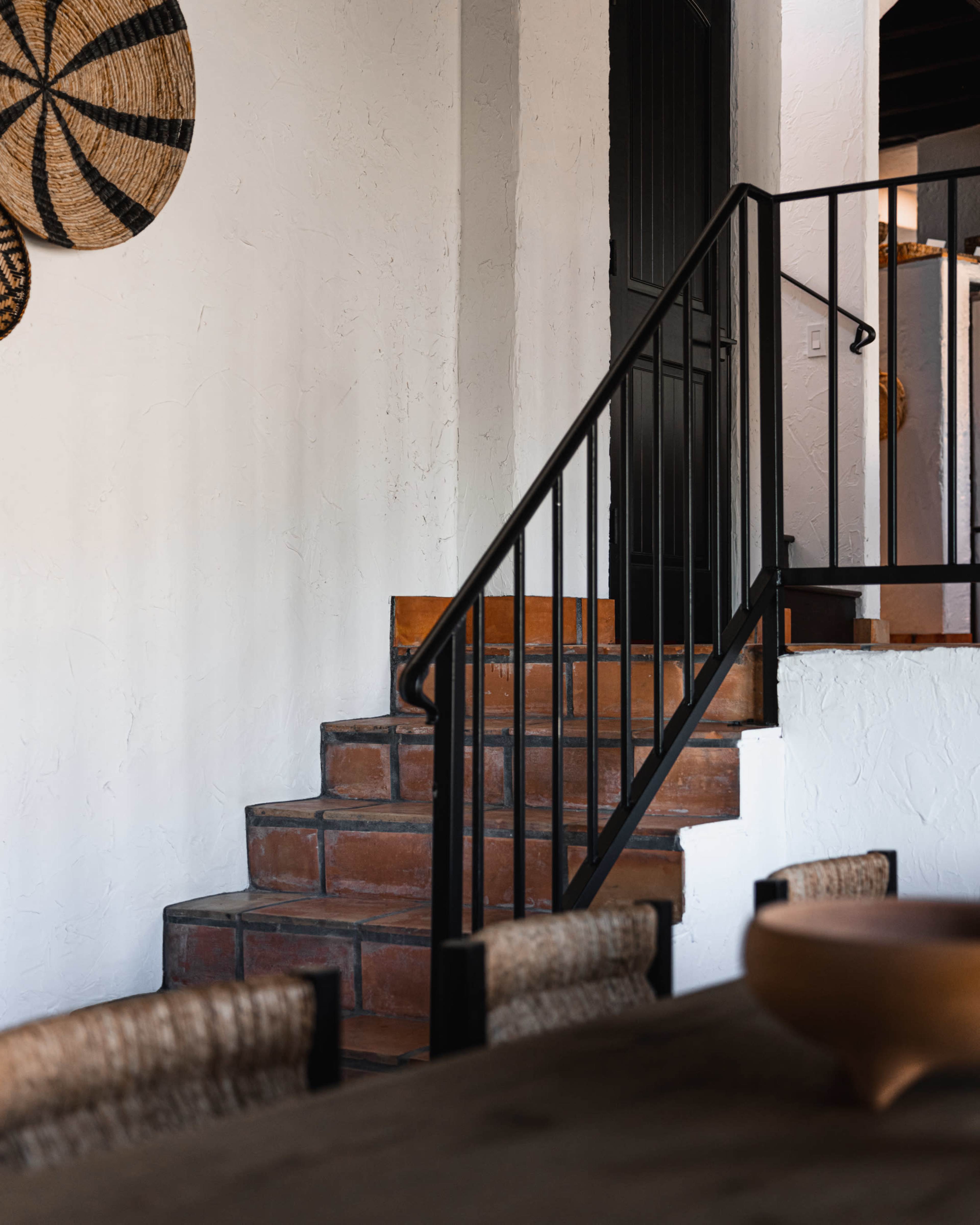 The image shows a staircase with terracotta steps leading to a black door, surrounded by white walls and woven wall decor.