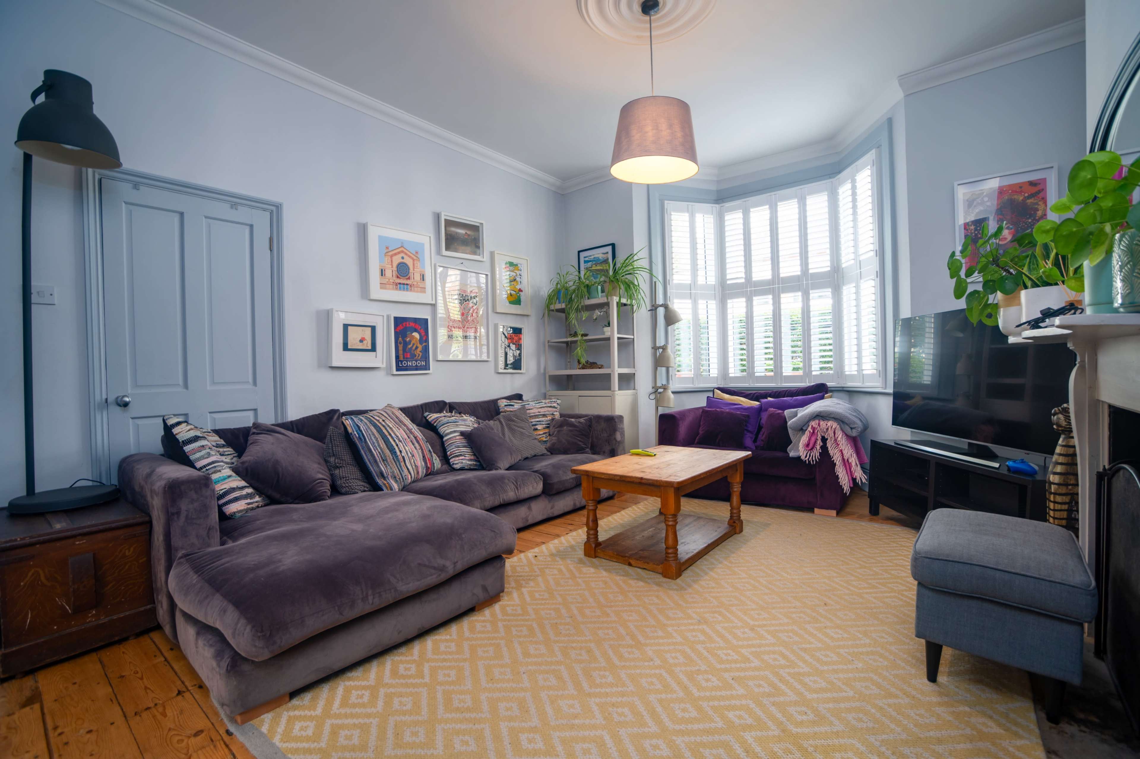The living room features a large gray sectional sofa, a wooden coffee table, and a television, with light coming through bay windows adorned with white shutters.