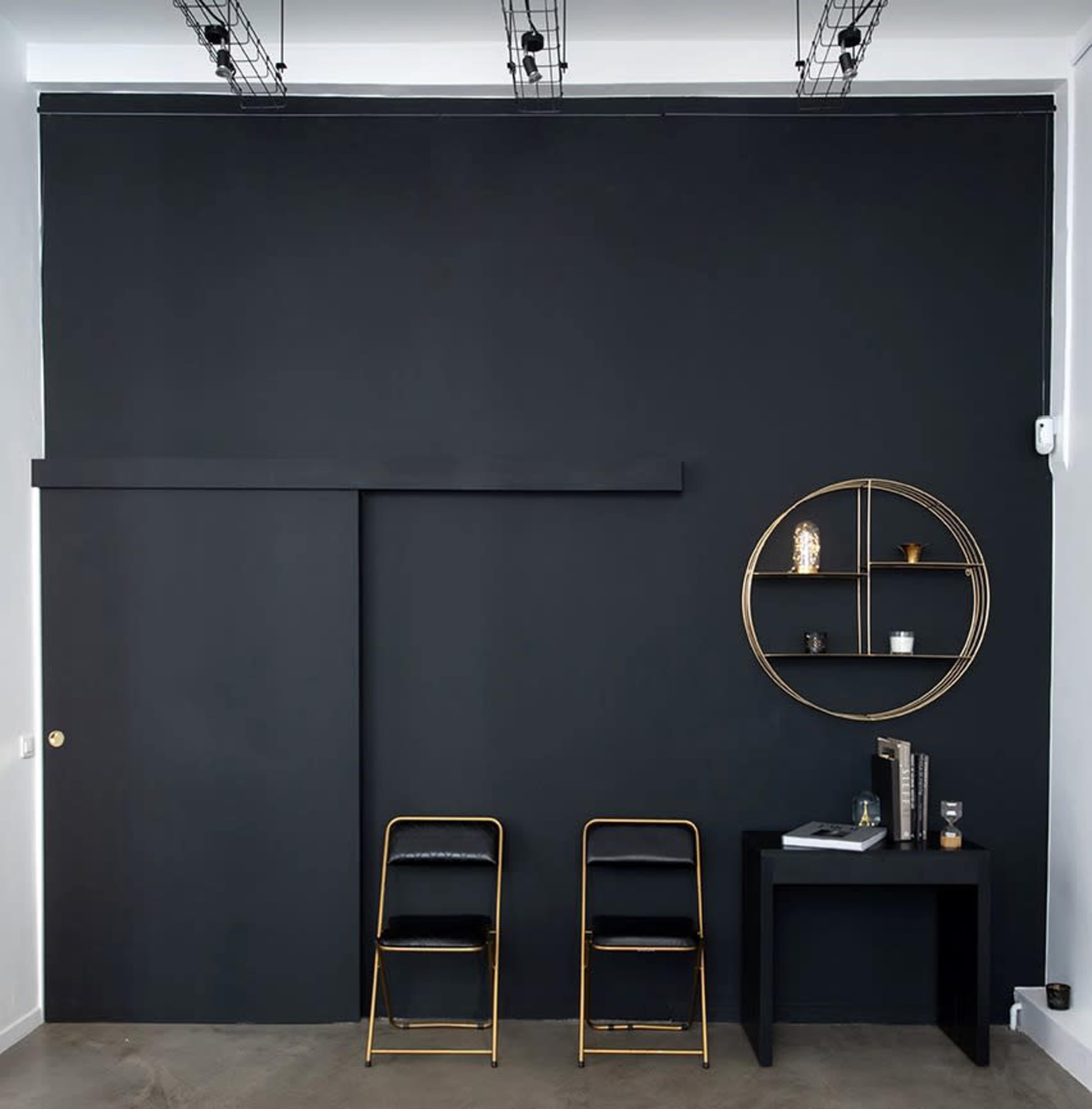 A minimalist interior with a black wall, a sliding door, two gold-framed chairs, and a circular shelf mounted on the wall.