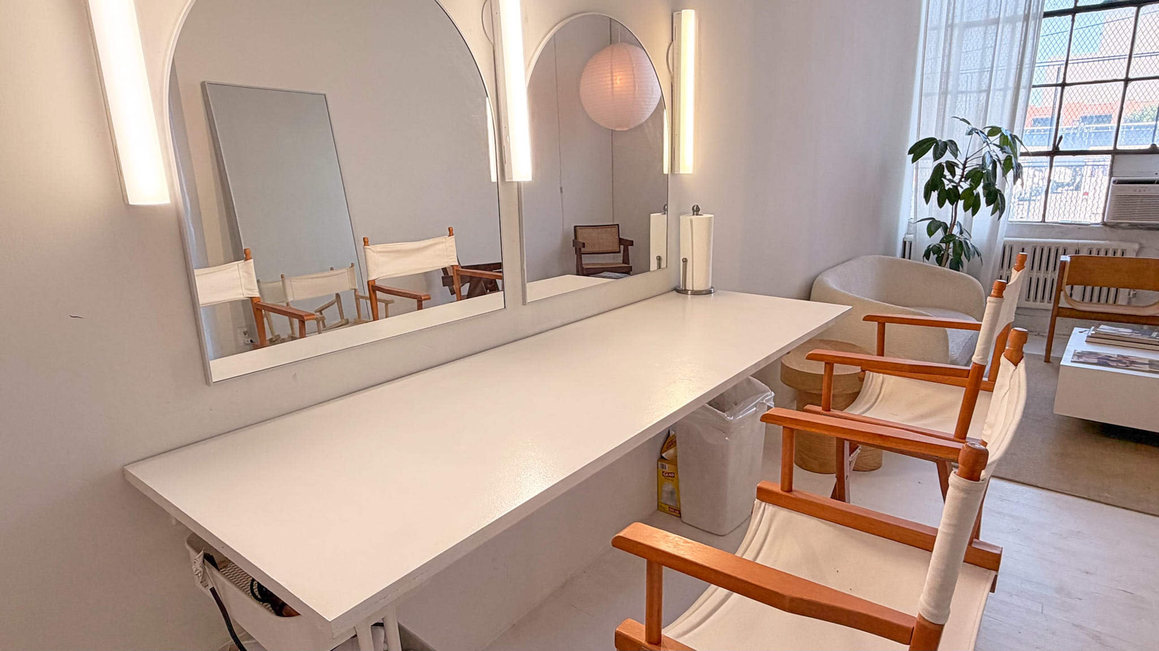 The image shows a bright, minimalist makeup station with two wooden chairs, a long white tabletop, and a large mirror reflecting natural light.