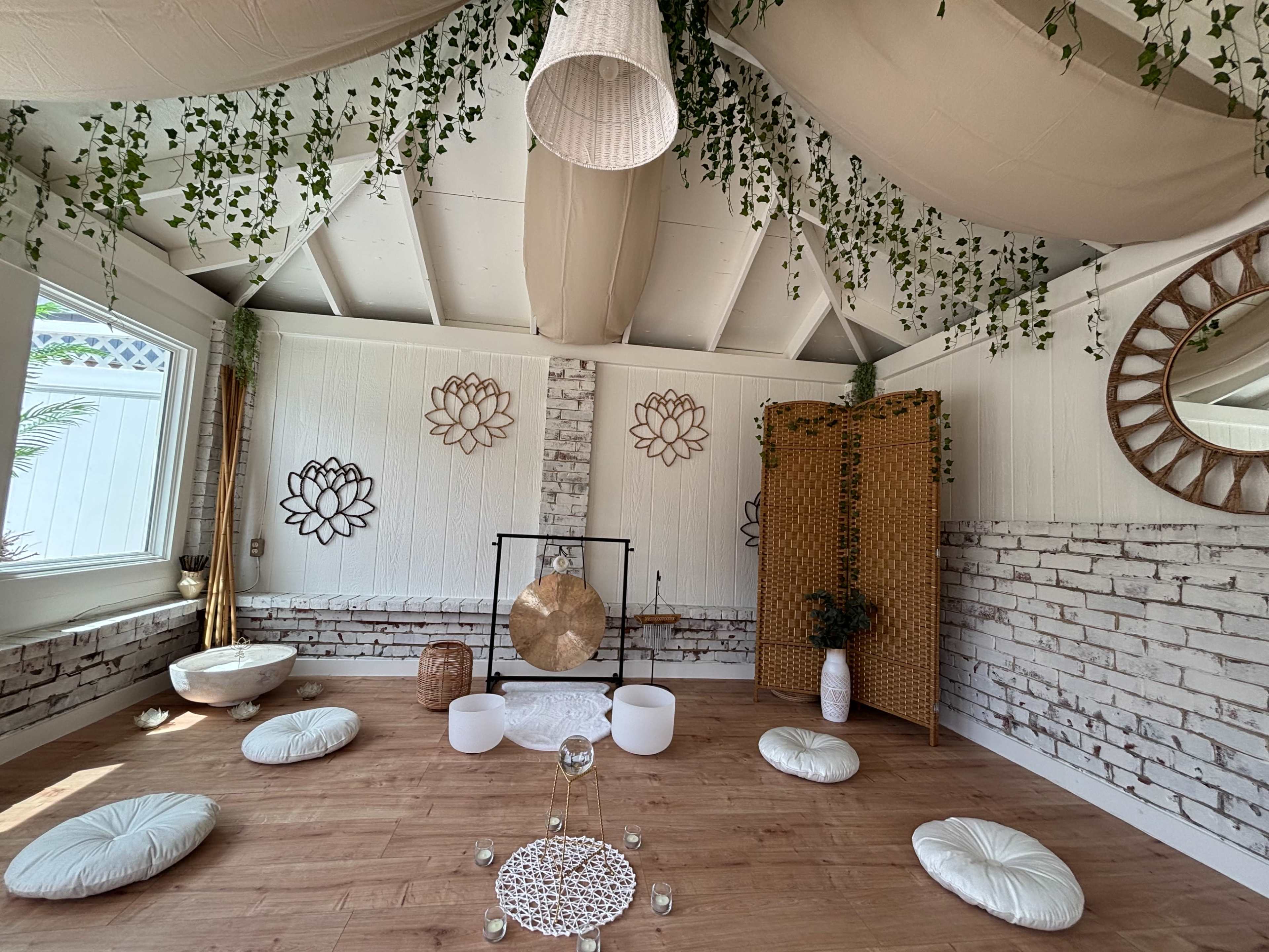 The image shows a tranquil interior space with wooden flooring, round cushions, a gong, and decorative elements including wall art and a folding screen.