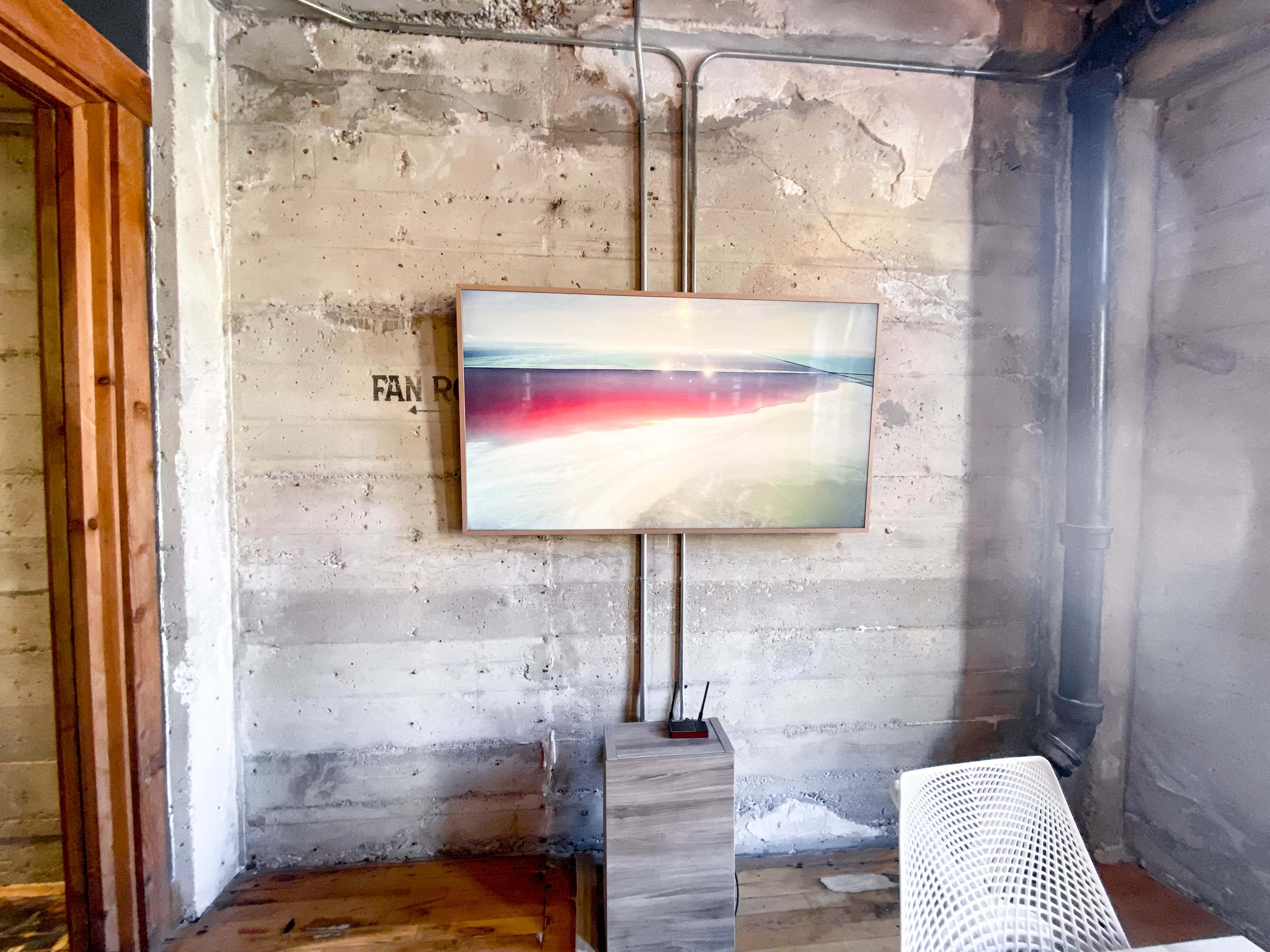A framed artwork is mounted on a concrete wall, with visible electrical conduits running alongside it, and a small wooden pedestal next to a white chair.