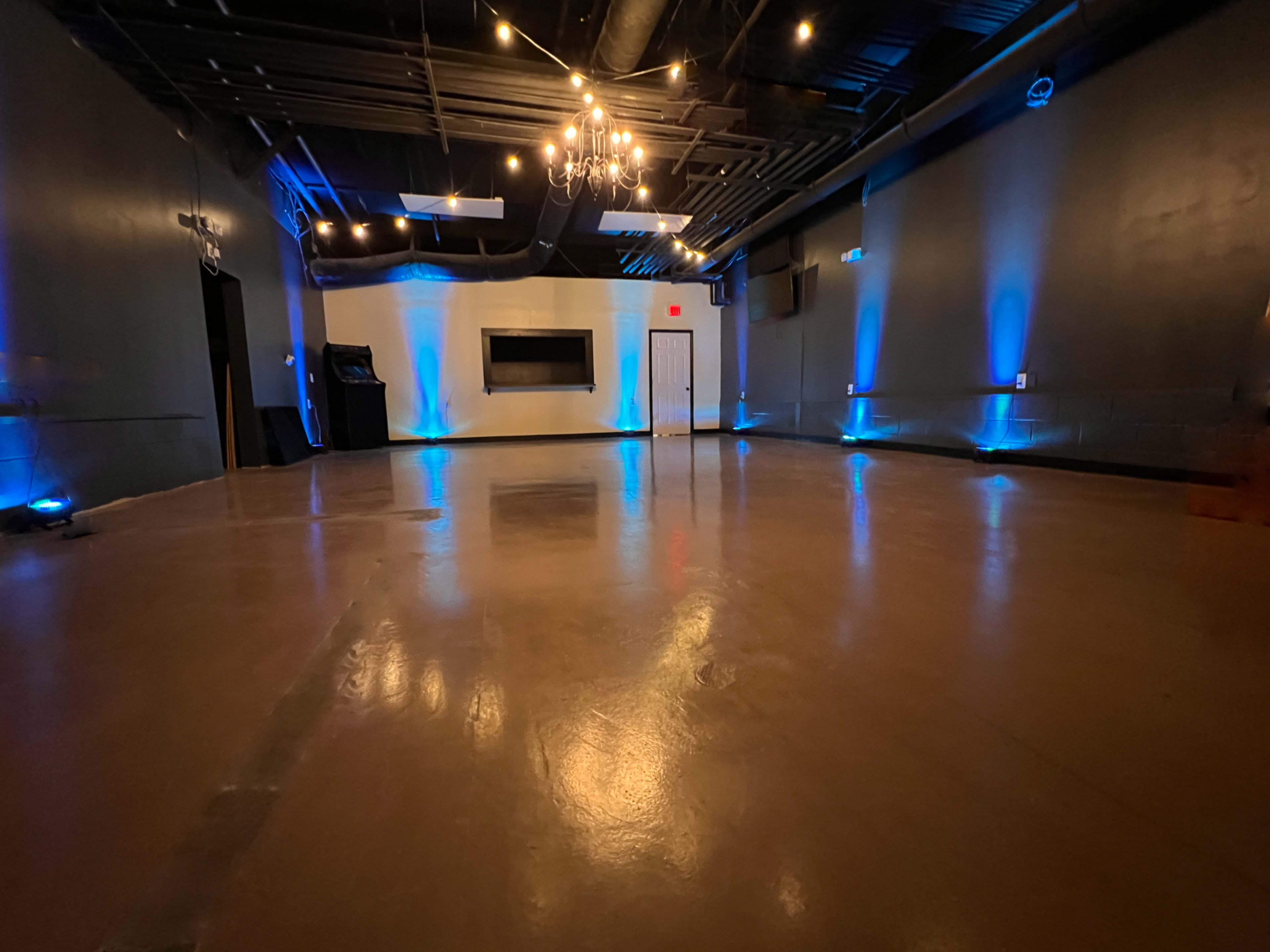 A spacious, empty event room with polished floors, blue lighting, and a chandelier.