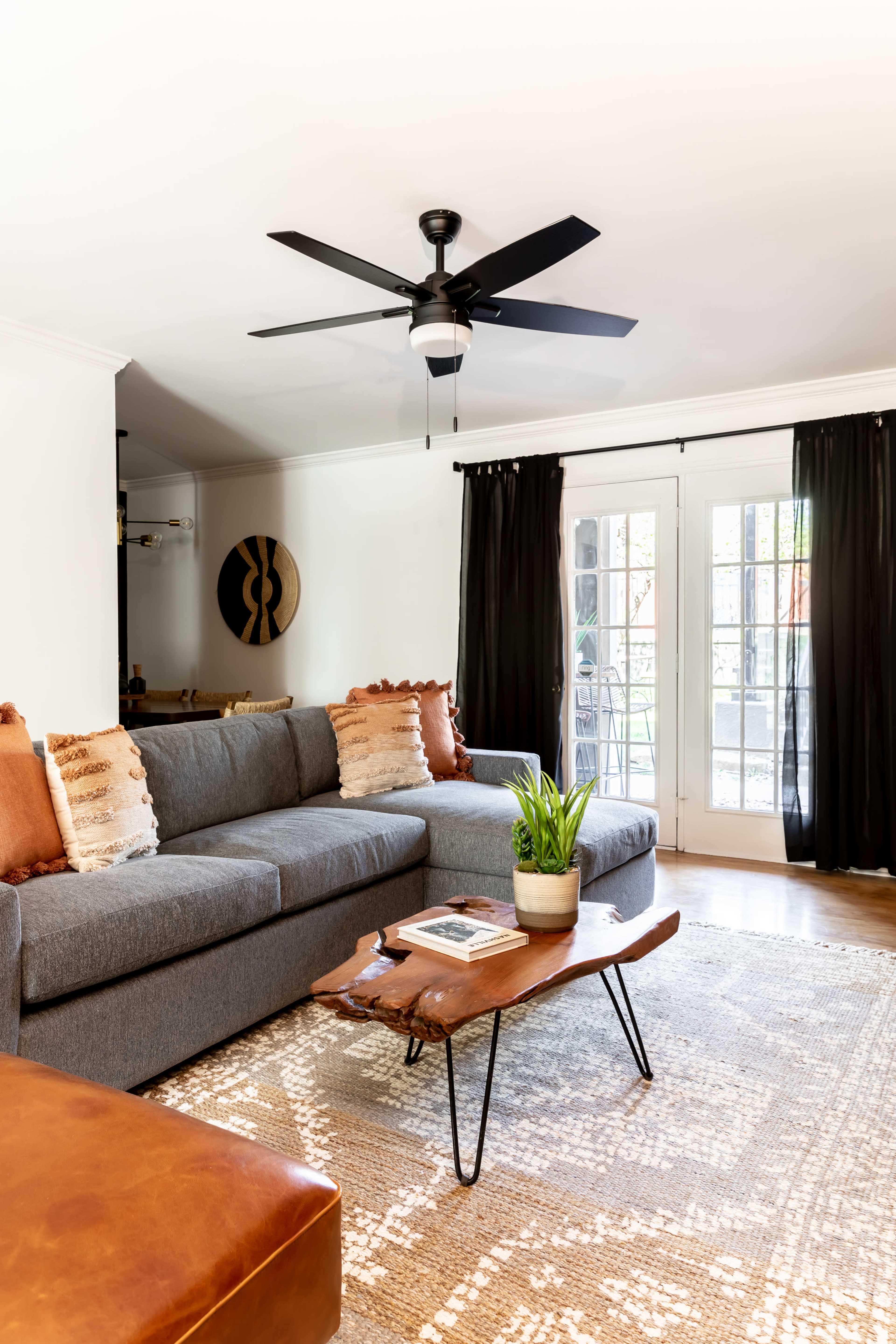 A modern living room features a gray sectional sofa, a wooden coffee table, and large windows covered with black curtains.