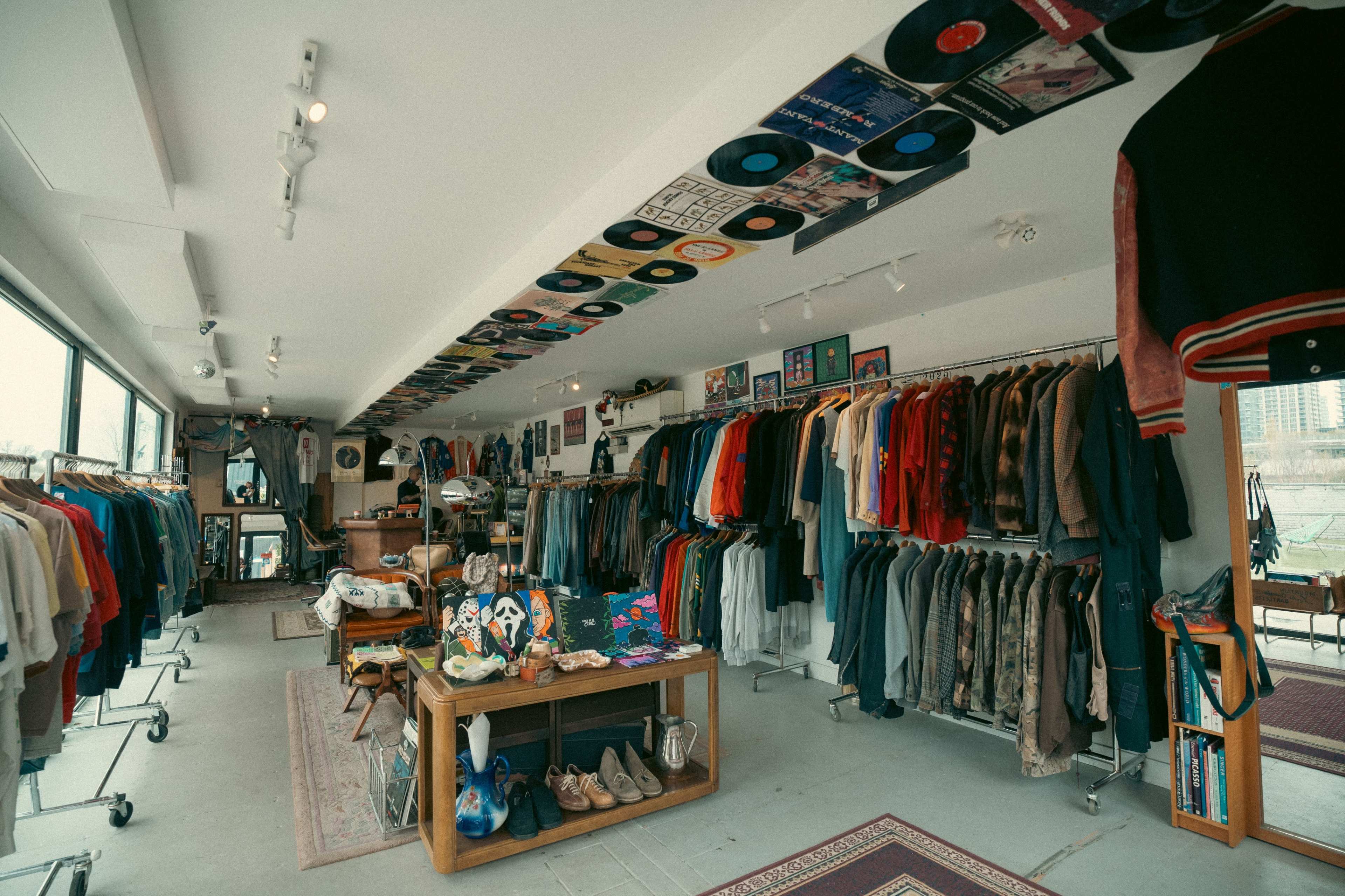 The image shows an inside view of a clothing store with racks of vintage apparel, a display table featuring accessories, and vinyl records adorning the ceiling.