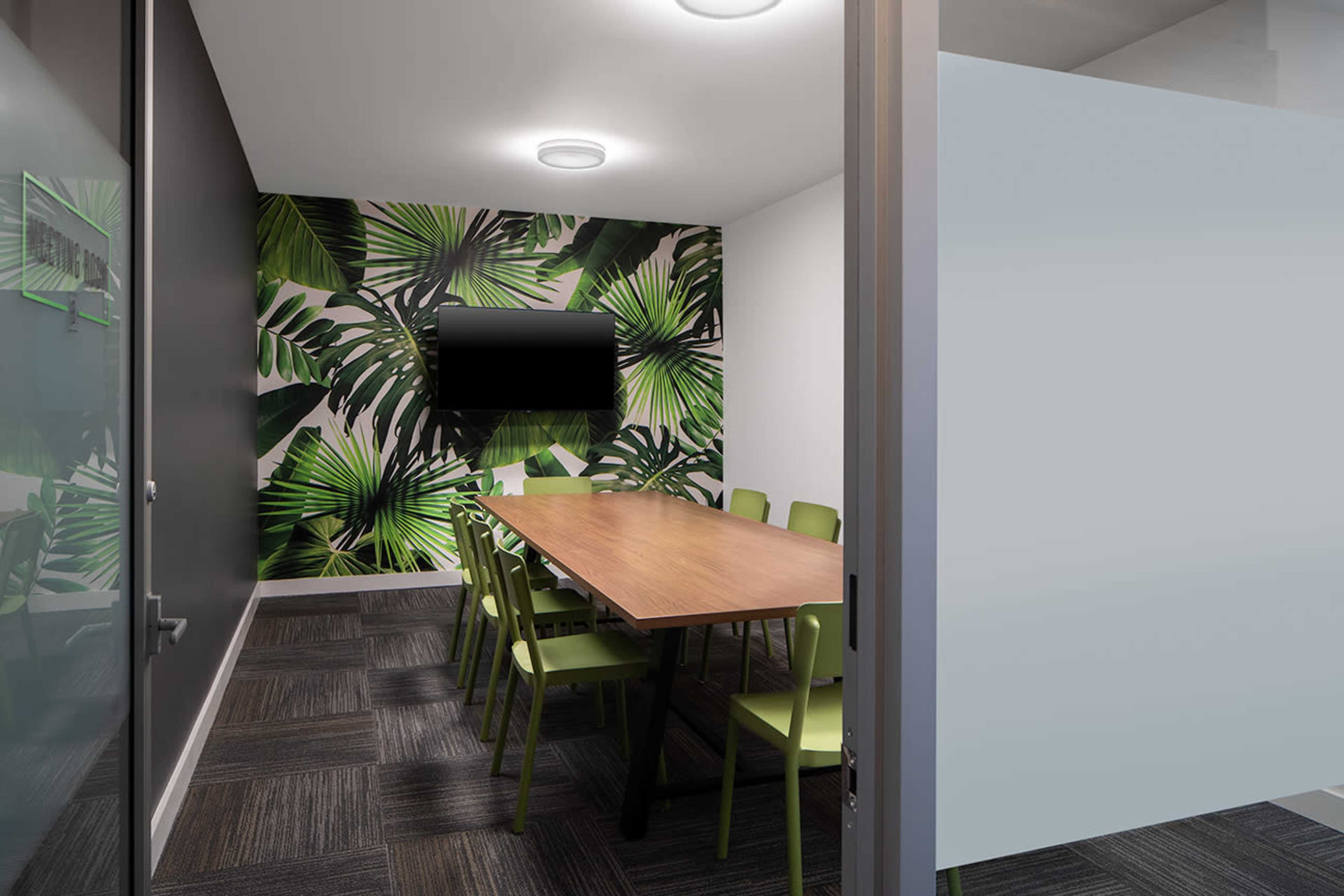 A conference room features a long wooden table surrounded by green chairs, with a wall decorated in tropical leaf patterns and a wall-mounted TV.