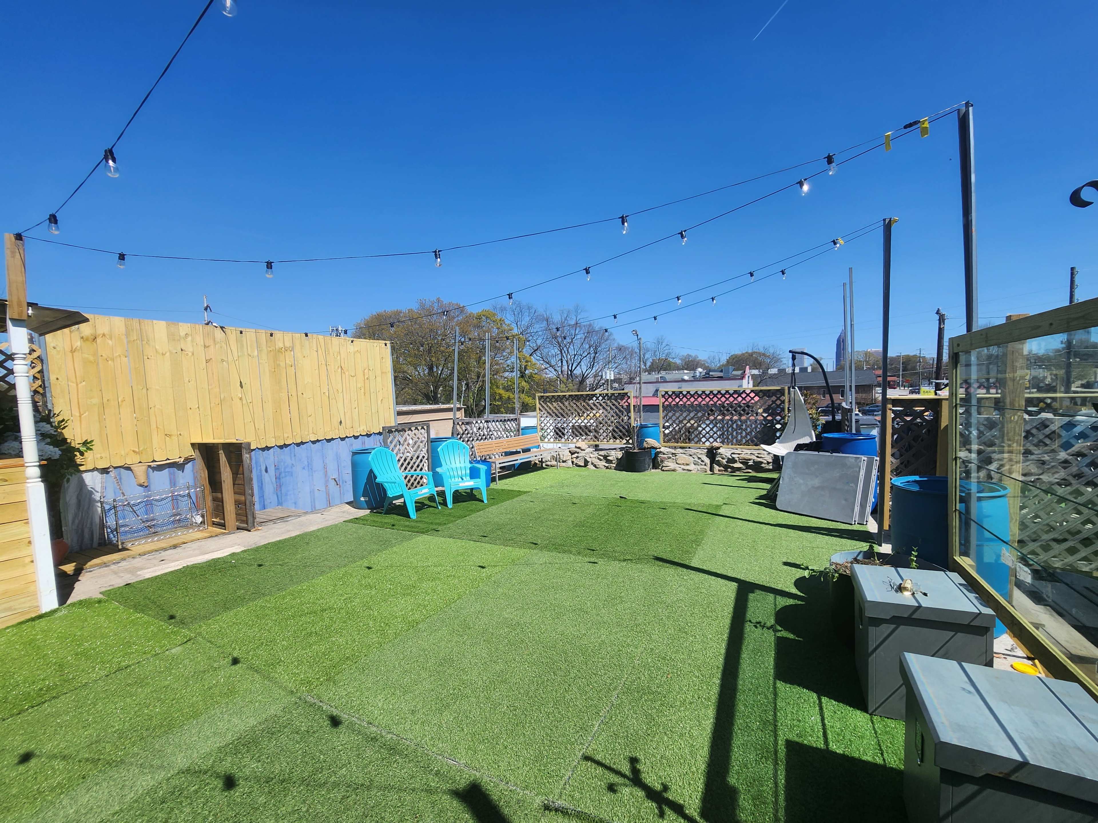 A rooftop terrace with artificial grass, two blue chairs, and string lights overhead against a clear blue sky.