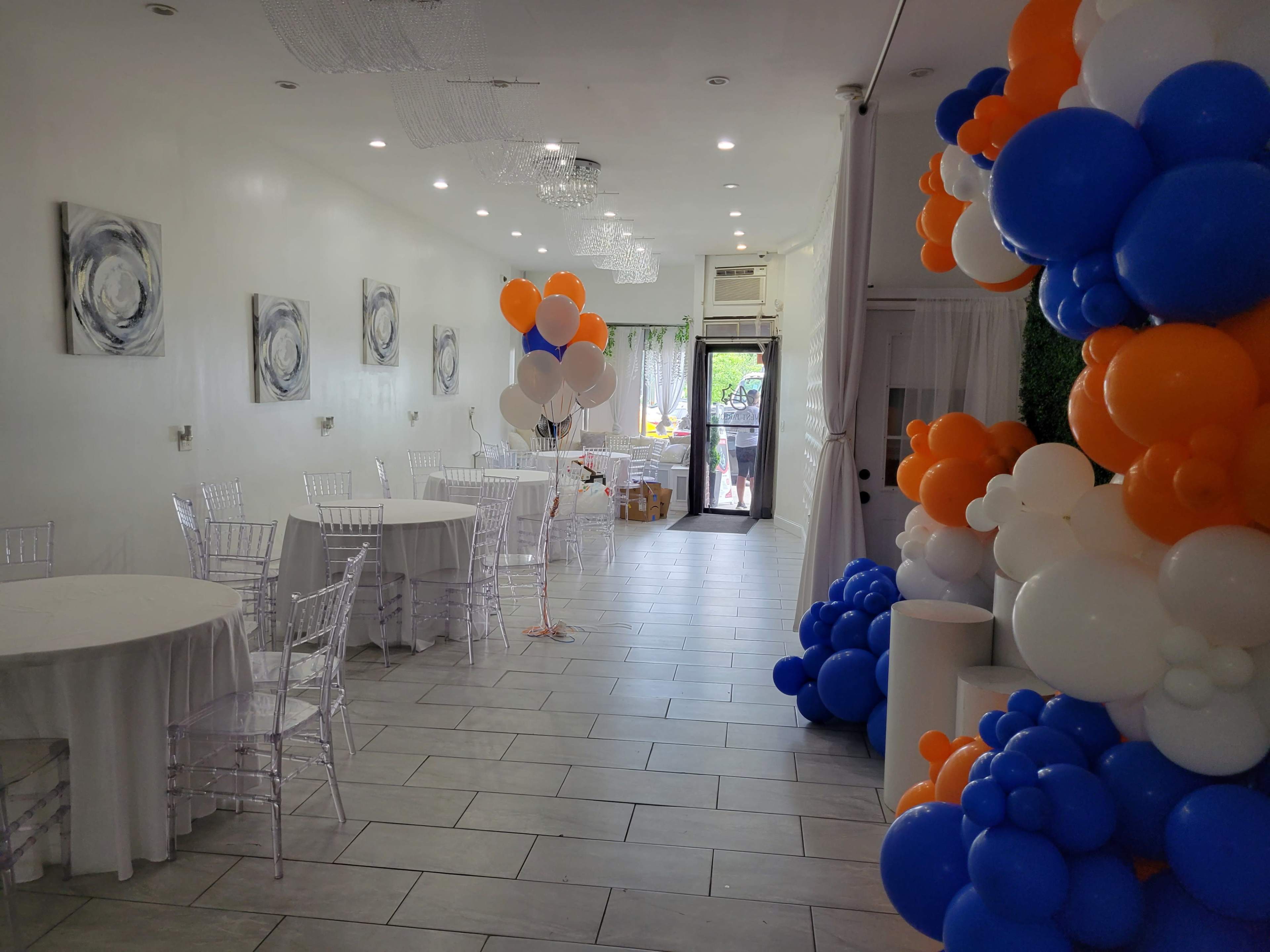 The image shows a decorated event space with white tables and a balloon arch in orange and blue colors.