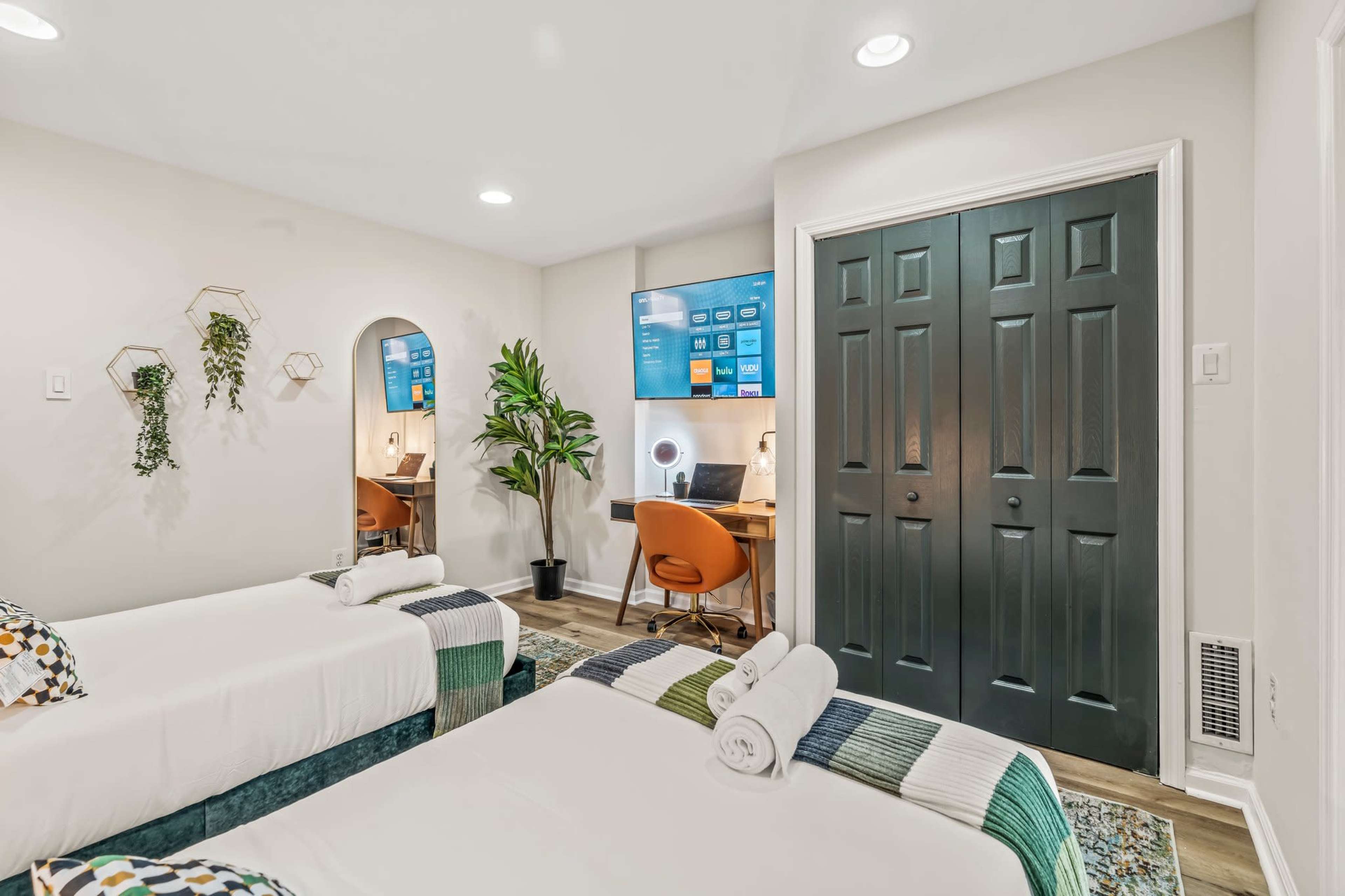 A room with two beds, a desk area with a computer setup, and a closet door, all arranged in a modern interior.