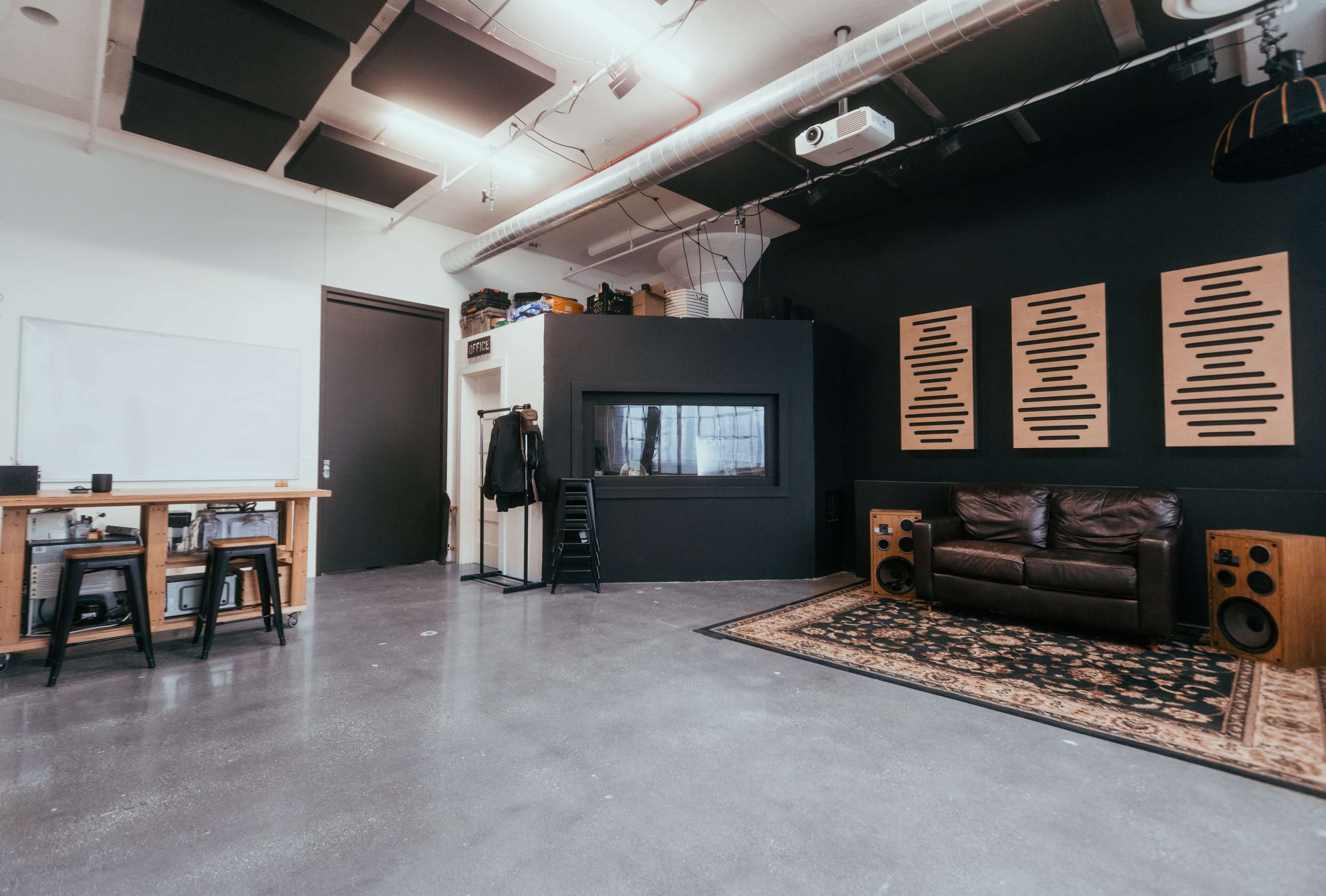 The image shows a modern studio space featuring a black wall with sound panels, a leather couch, and audio speaker equipment, alongside a whiteboard and a table with stools.
