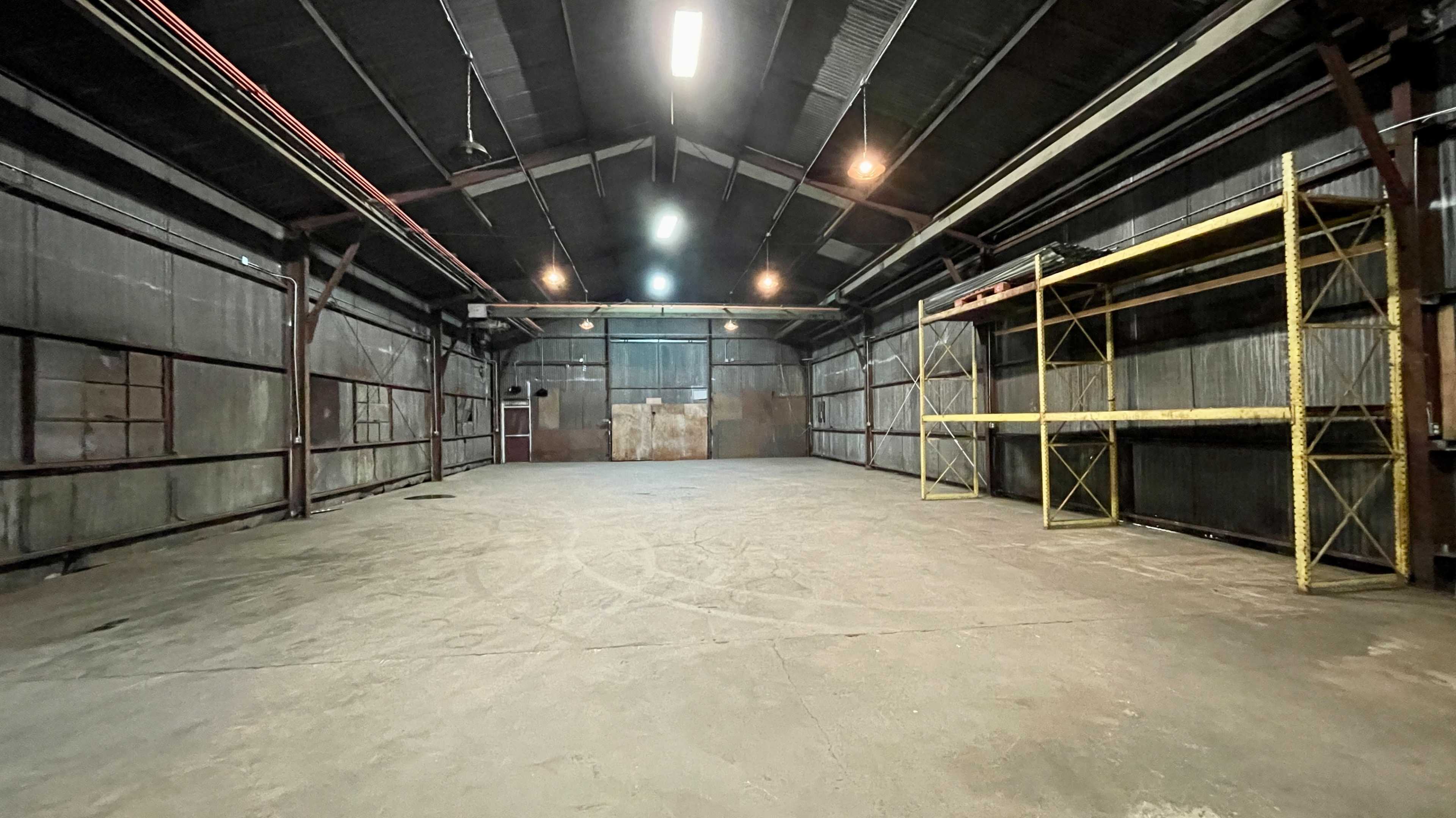 A spacious, empty warehouse with metal walls, concrete flooring, and a single row of yellow shelving along one side.