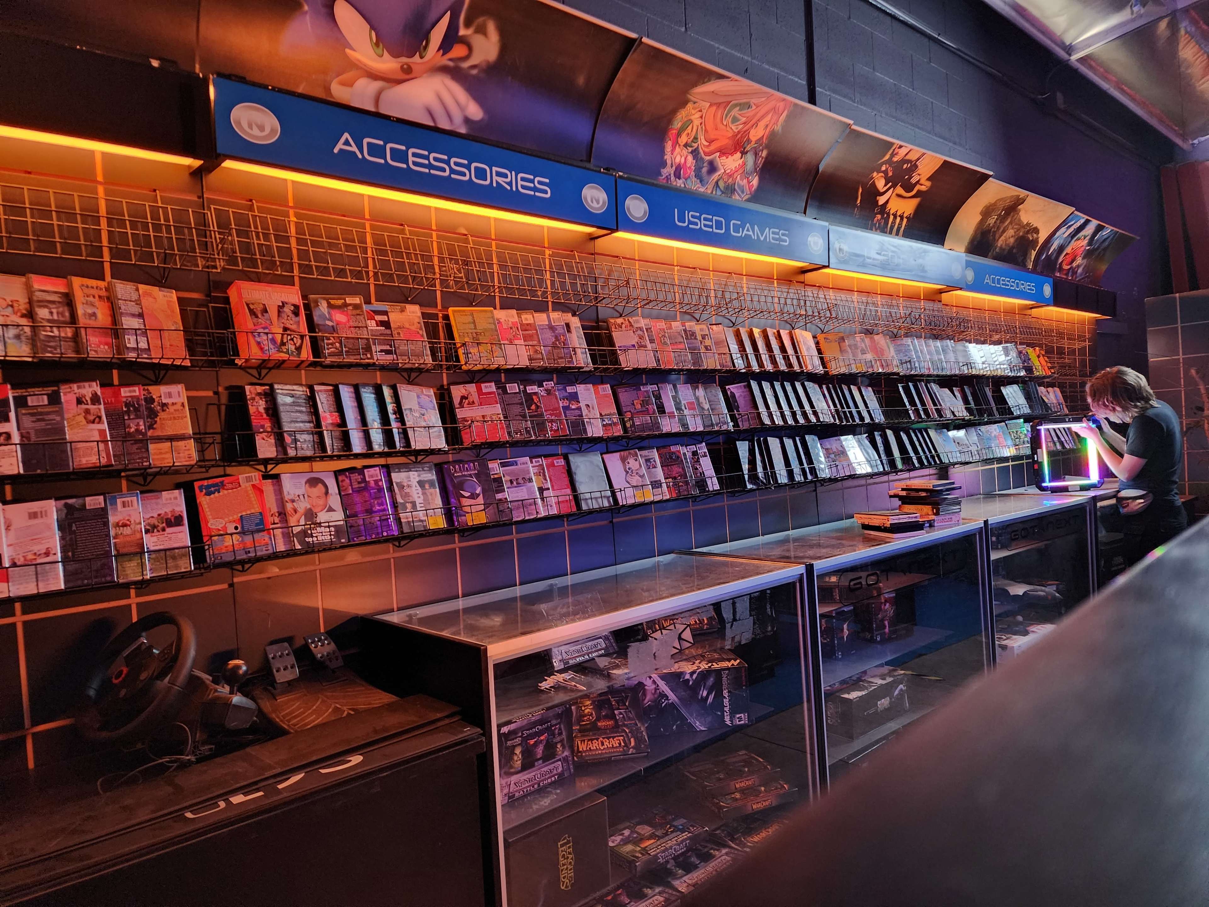 The image shows a video game store with shelves organized full of games and accessories, highlighted by orange lighting, while a person is seen working at a counter.