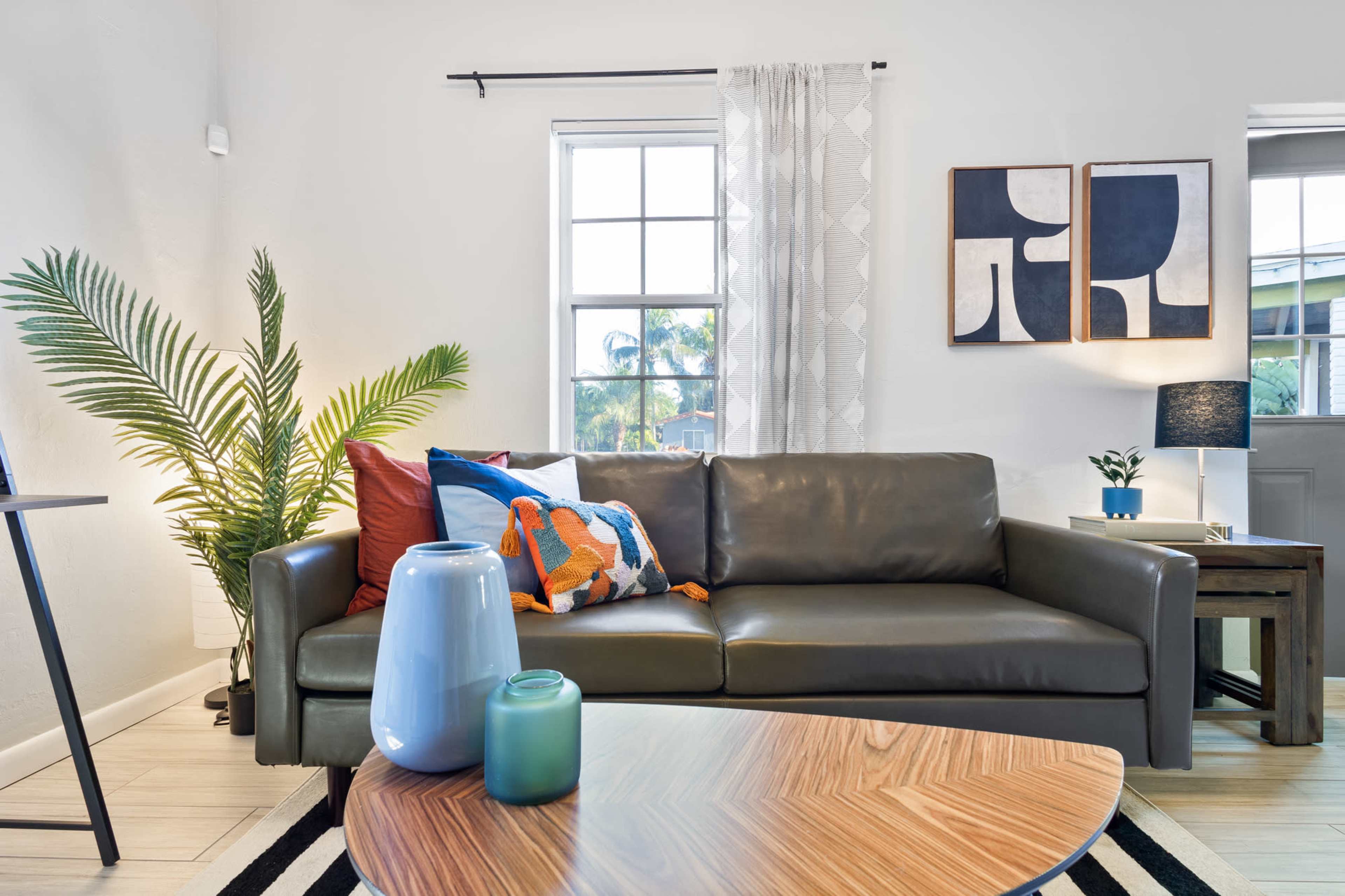 A living room with a grey leather sofa adorned with colorful pillows, a wooden coffee table, and decorative plants next to a window.