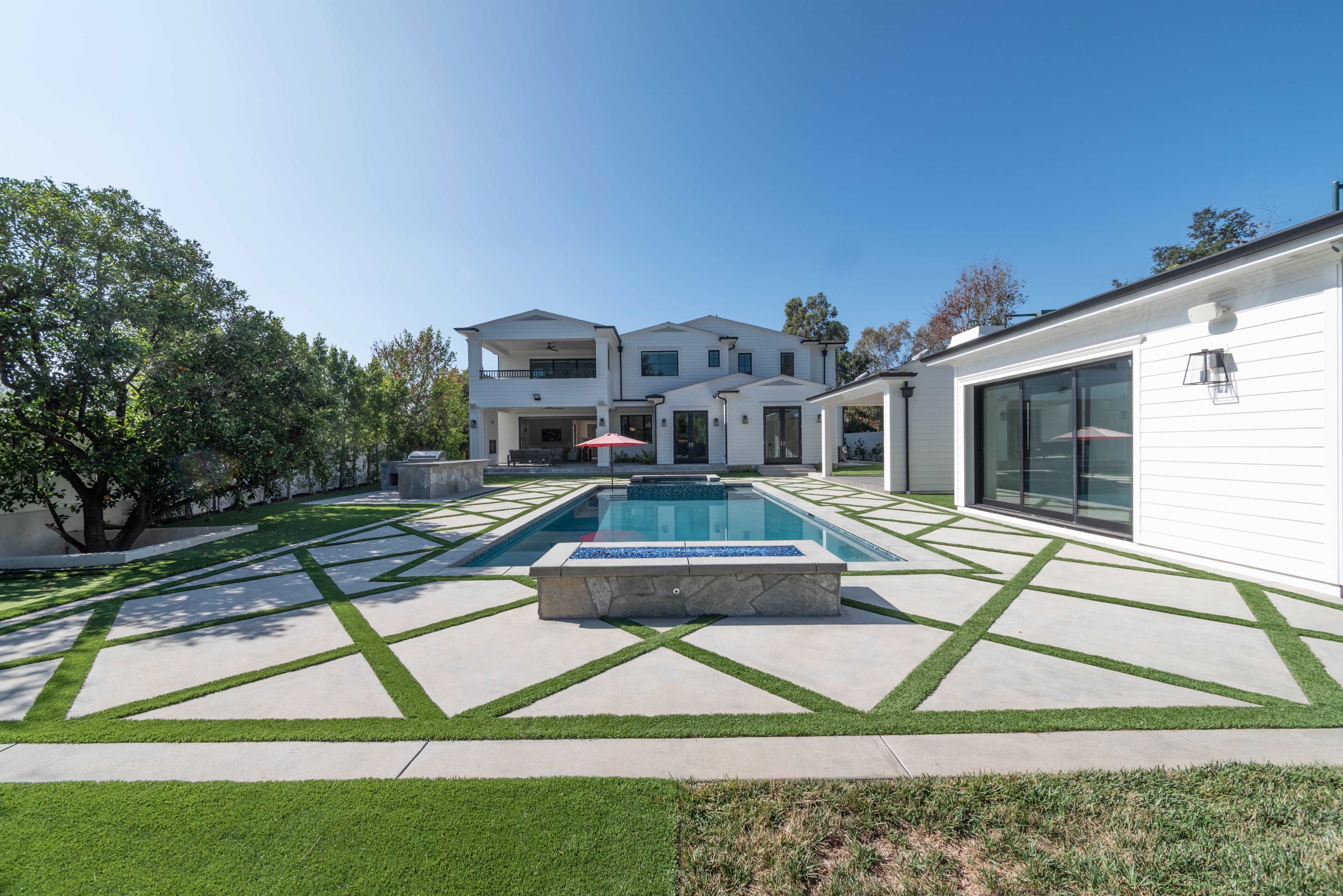 A modern two-story house with a swimming pool is surrounded by neatly patterned concrete and artificial grass landscaping.