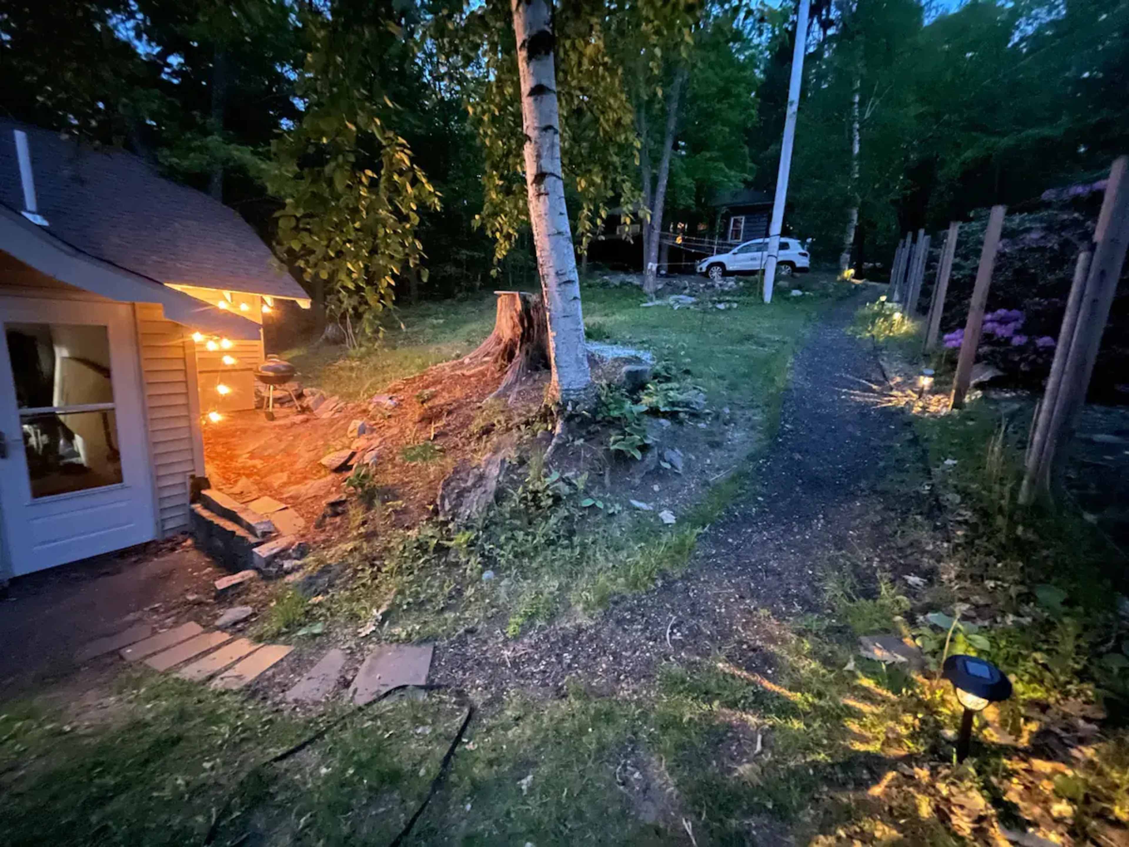 A path winds through a wooded area, with a tree stump and a cabin illuminated by string lights on one side and a house visible in the distance on the other.