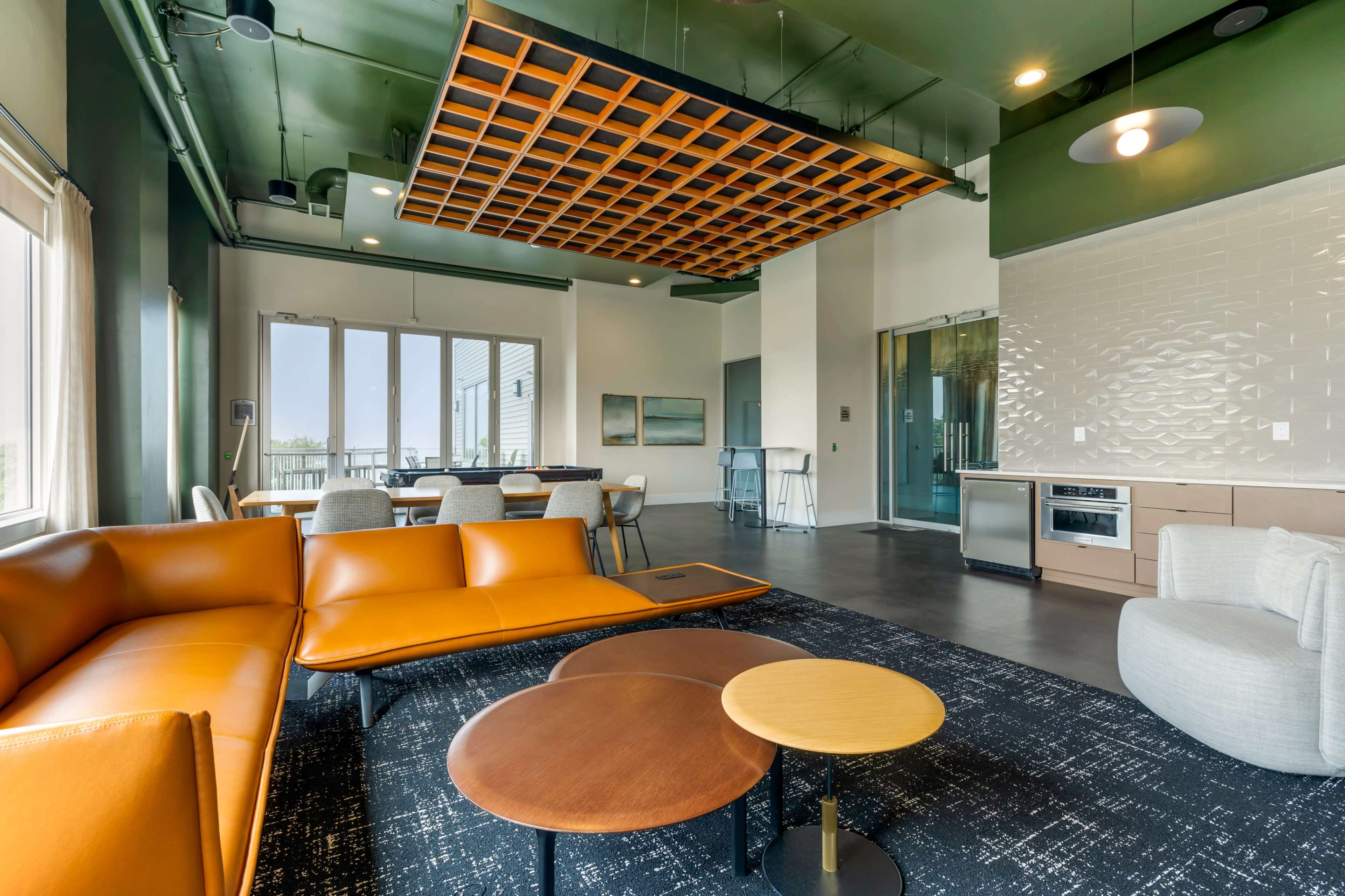 The image shows a modern lounge area featuring a brown leather sofa, two round coffee tables, and a wooden grid ceiling, with large windows allowing natural light to fill the space.