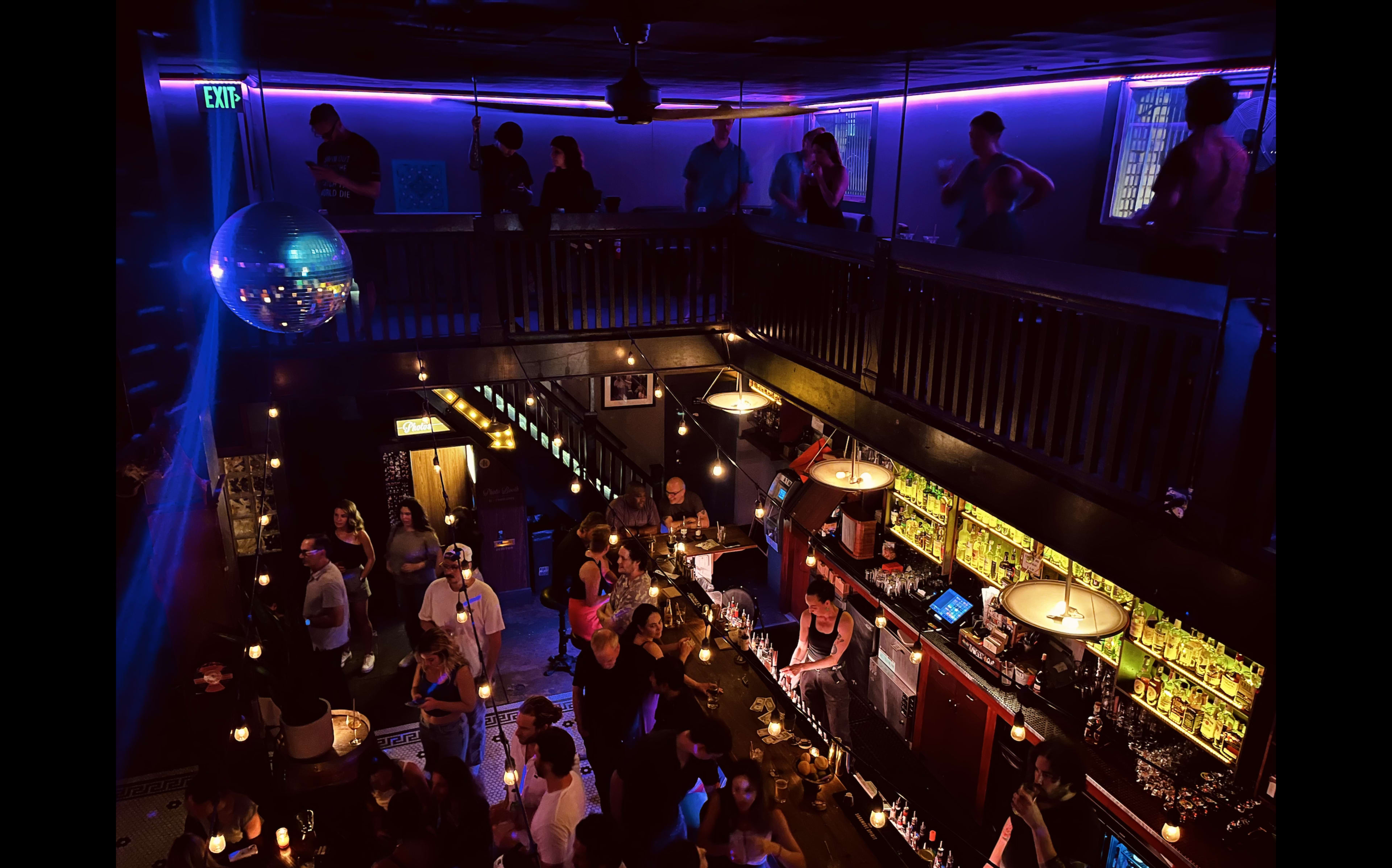 A lively bar scene fills the image, featuring people socializing both on the ground floor and the balcony above, illuminated by neon lights and a disco ball.