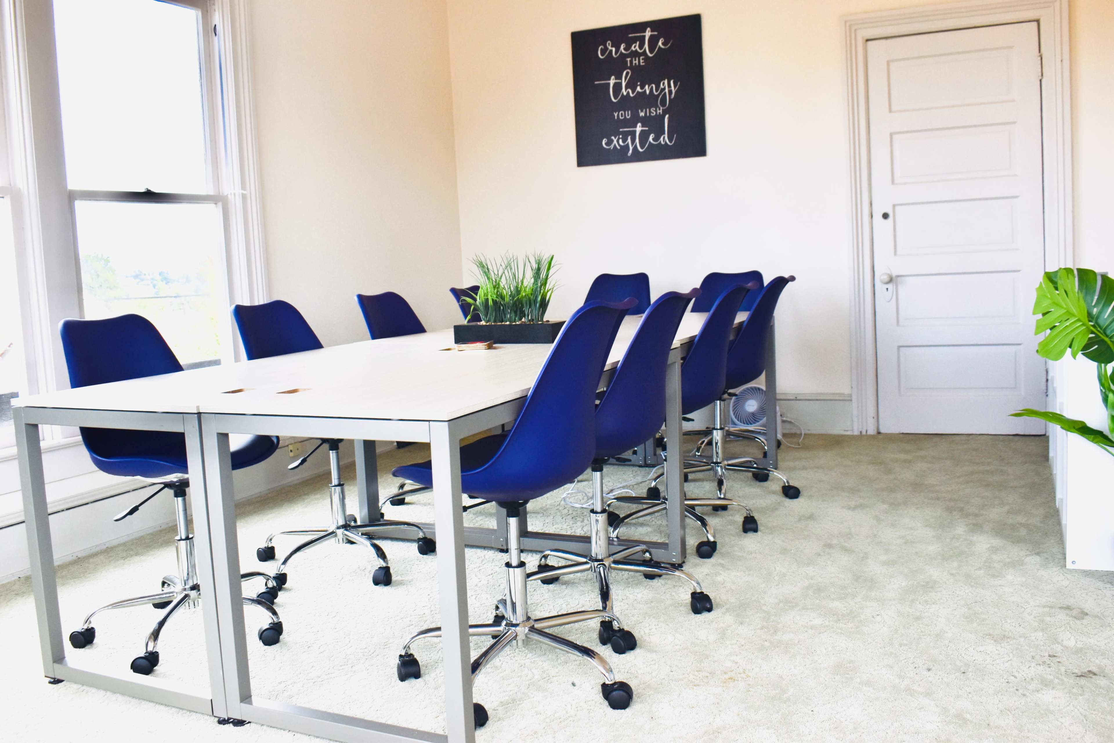 A conference room features a large white table surrounded by blue swivel chairs, with a potted plant and a motivational quote on the wall.