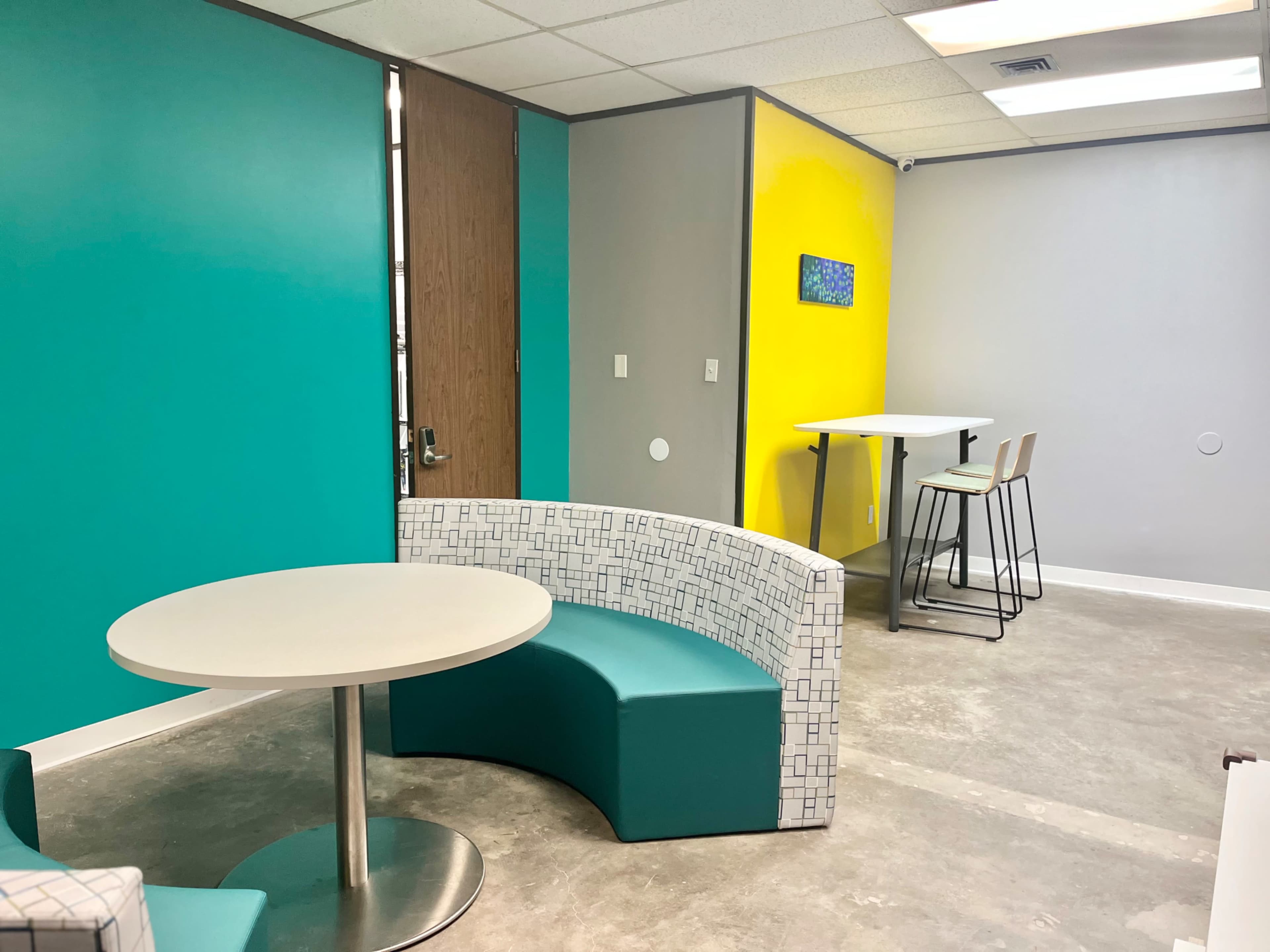 The image shows a modern interior space featuring a curved seating area with a white table, colorful walls in teal and yellow, and a high table with stools in a corner.
