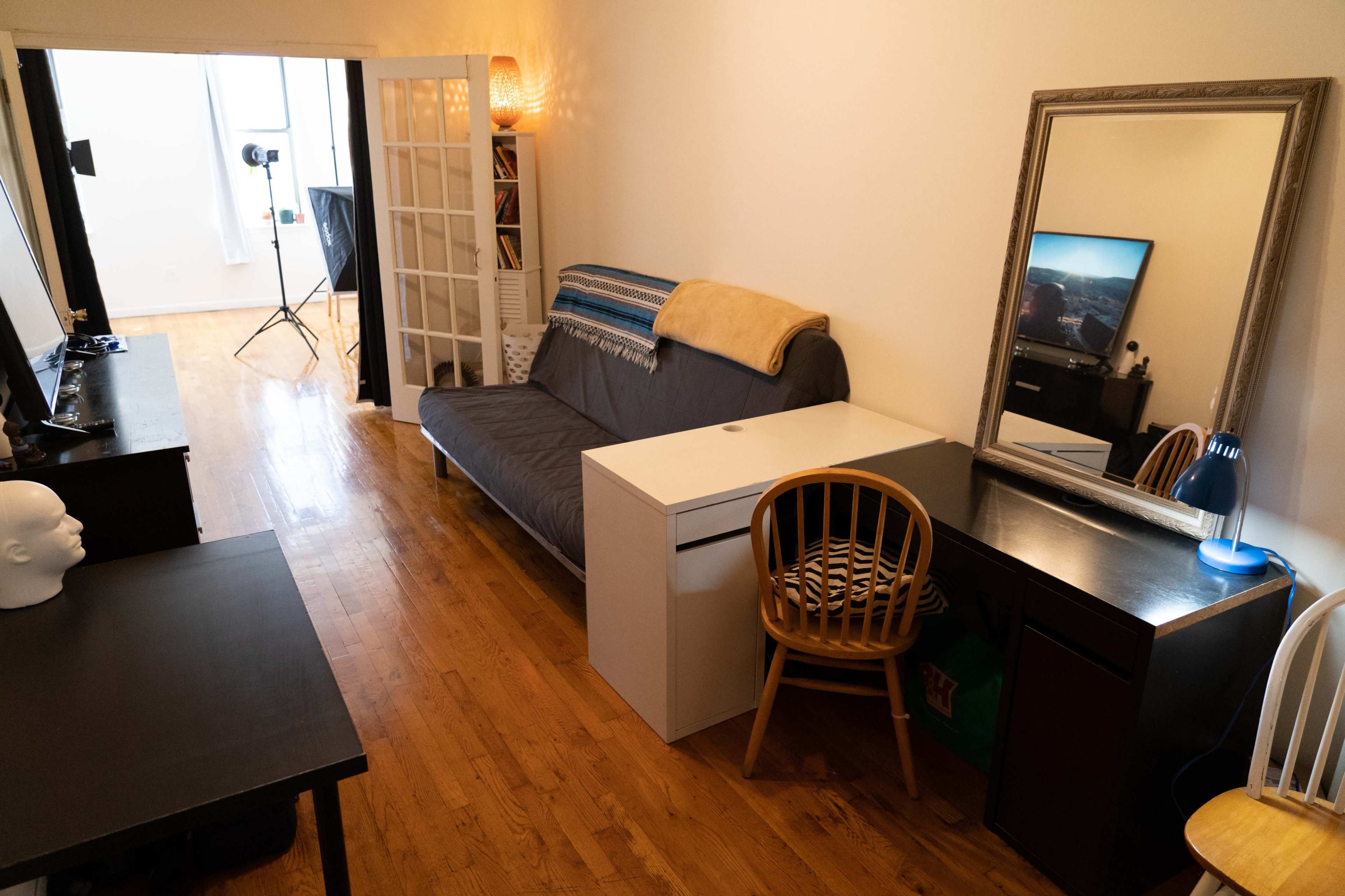A small living room with a futon, a desk with a lamp, and a large mirror against the wall, leading to a well-lit area in the back.