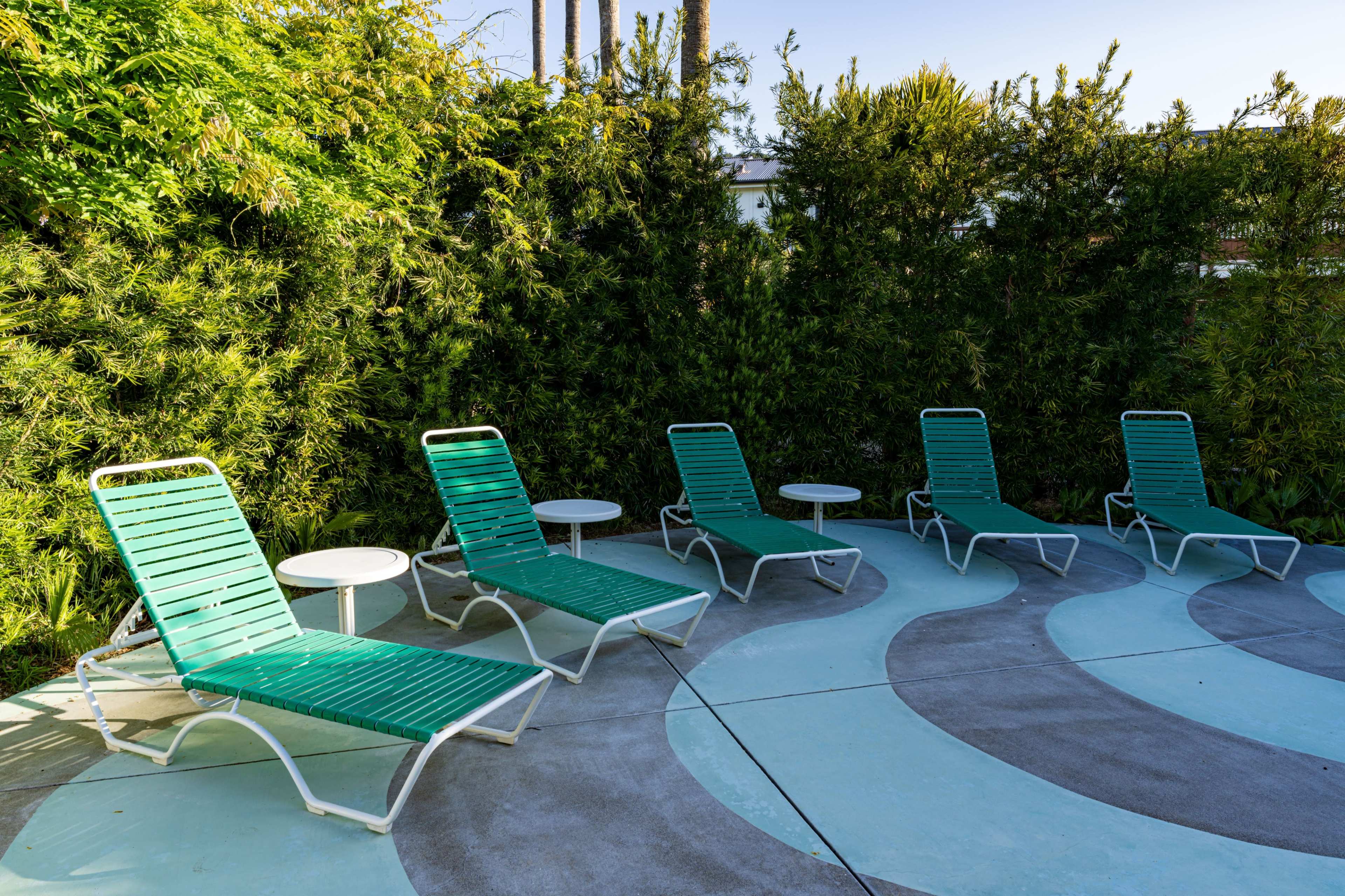 Five green lounge chairs with white side tables are arranged in a curved pattern on a concrete patio surrounded by lush greenery.
