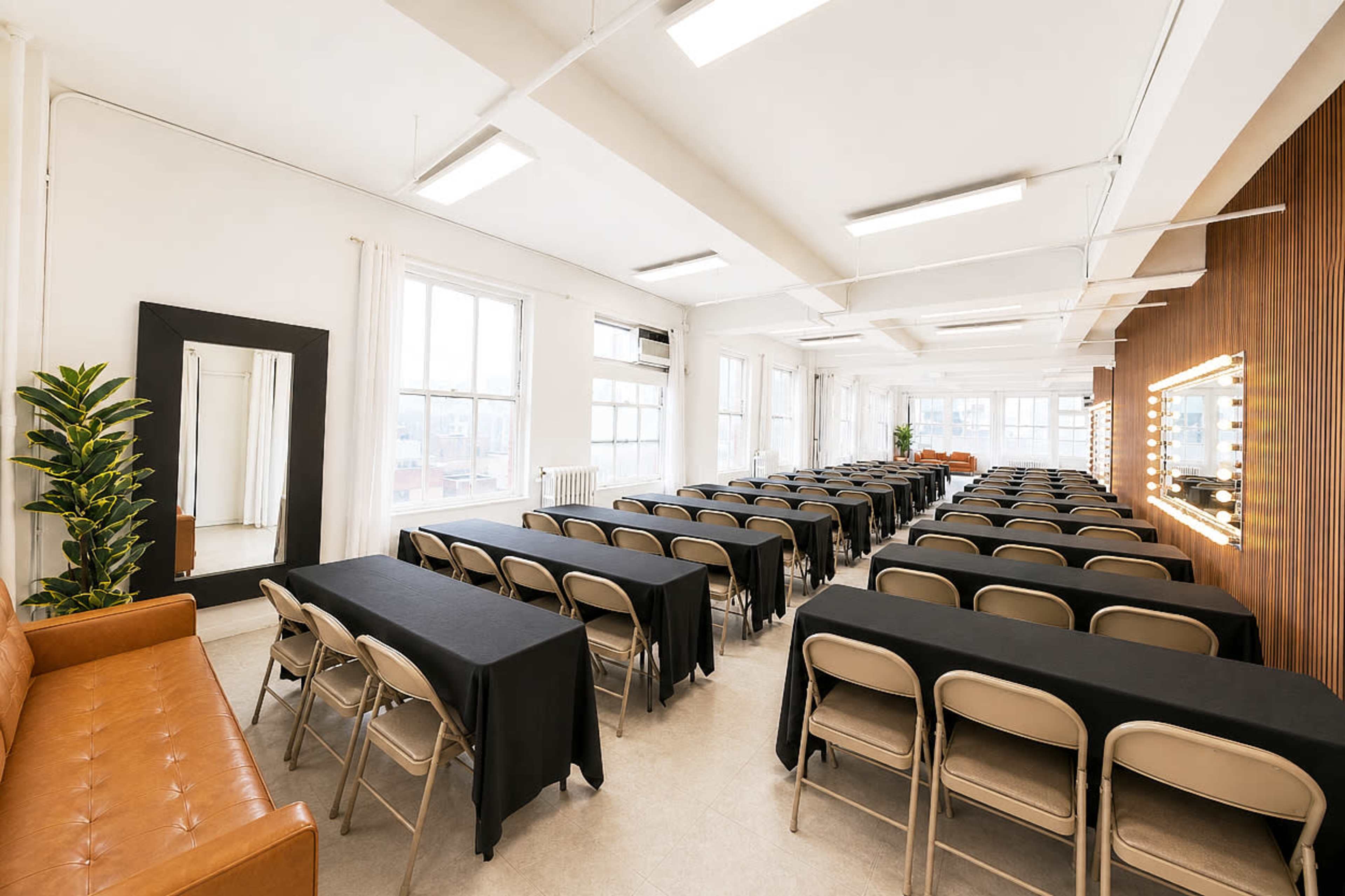 The image shows a spacious room arranged with rows of tables covered in black cloth and lined with folding chairs, with a brown sofa and a large mirror on one side.