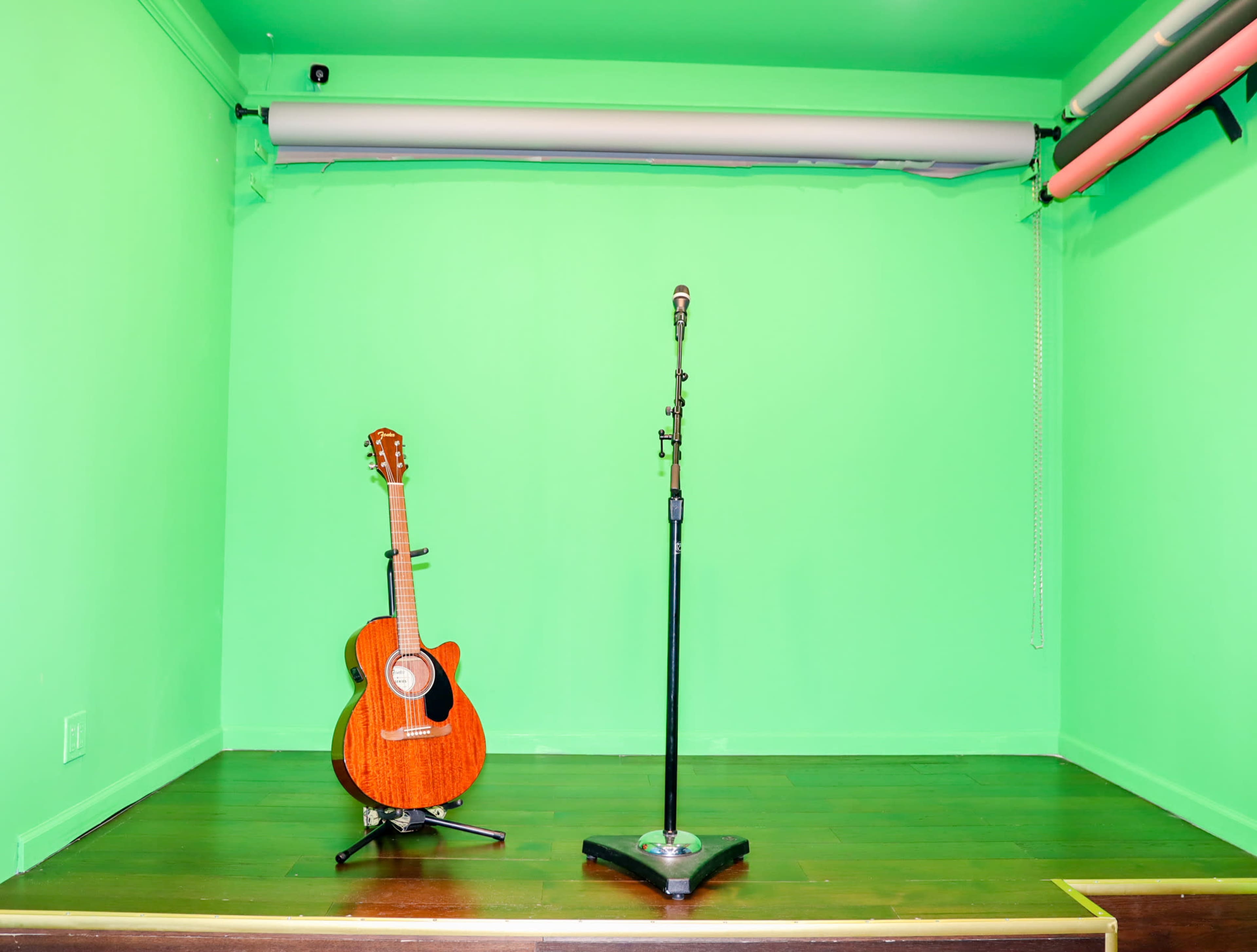 An empty stage with a brown acoustic guitar leaning against the wall and a microphone on a stand, all set in a green backdrop.