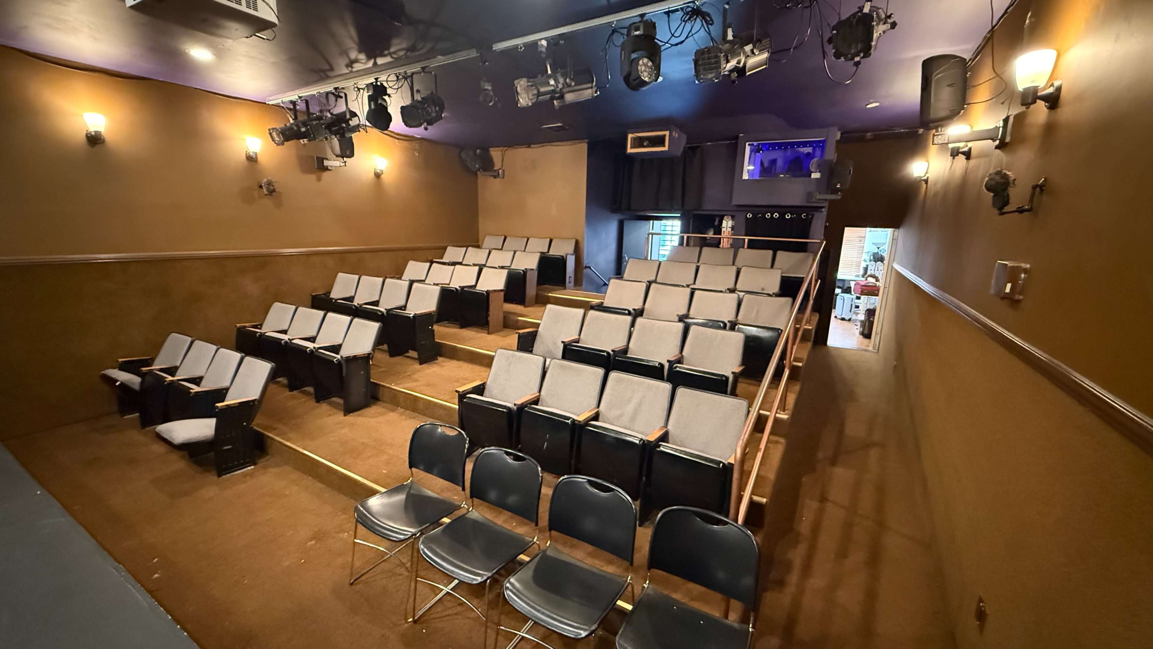 The image shows a small theater with tiered seating, spotlights, and a stage area in the background.