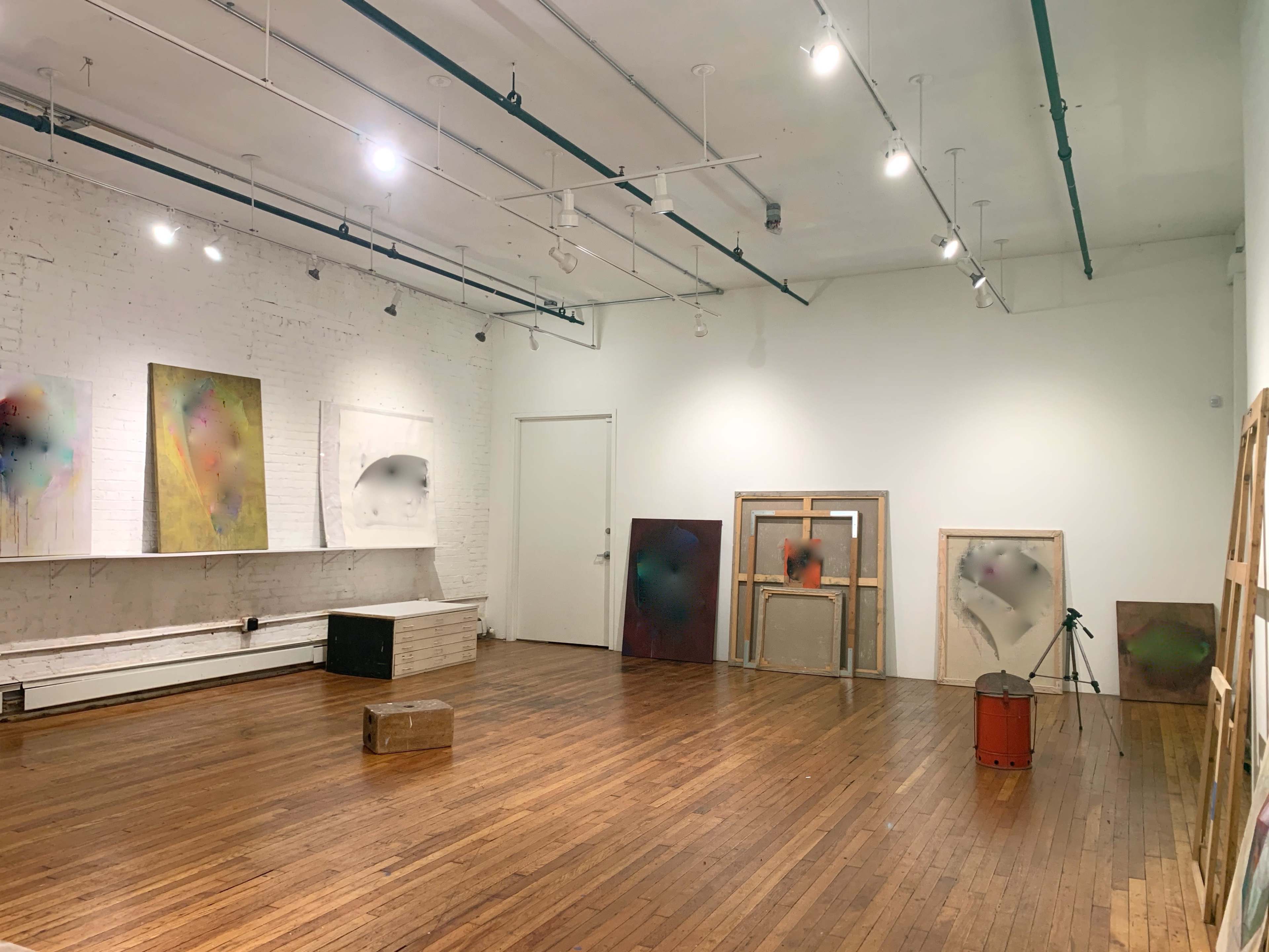 An art gallery with several abstract paintings displayed on white walls and some wooden frames leaning against a wall on a wooden floor.
