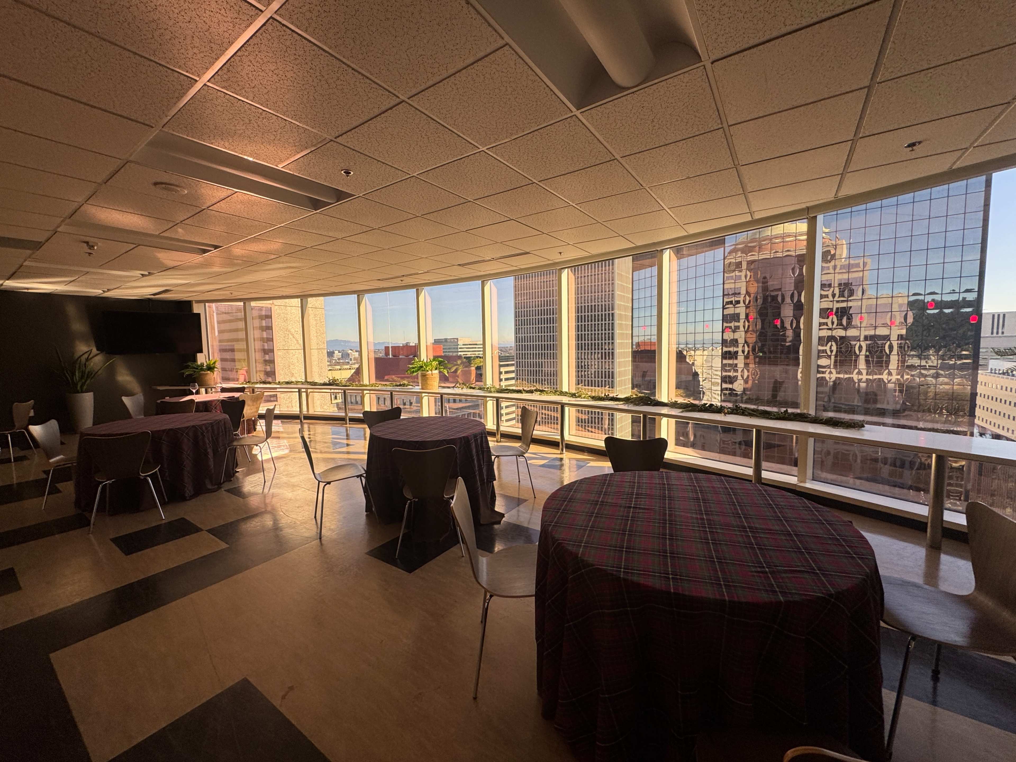 The image shows a spacious room with multiple tables covered in checkered tablecloths, having large windows that offer a view of city buildings.