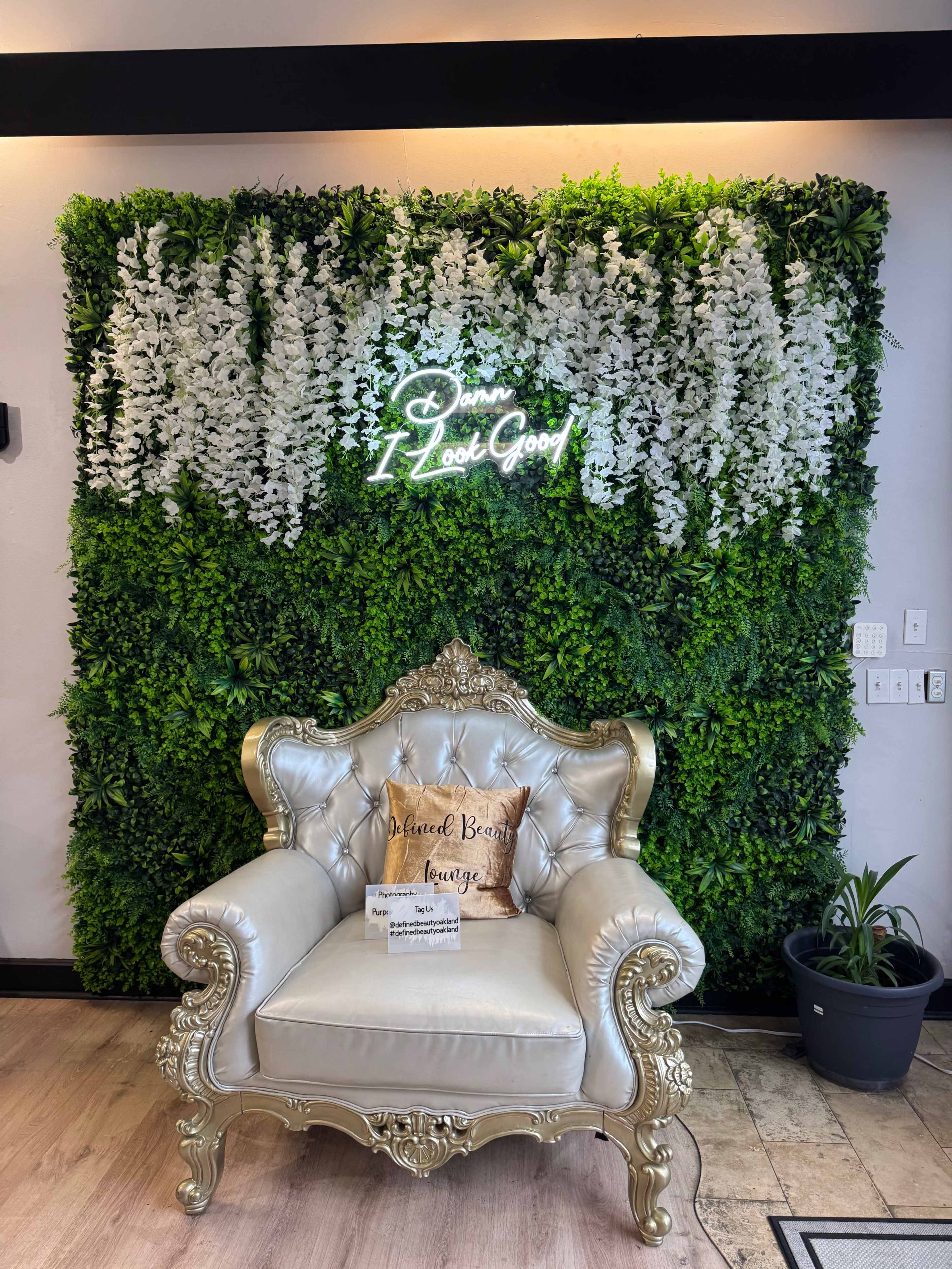 A luxurious silver tufted chair is positioned in front of a lush green wall adorned with white flowers and a glowing neon sign that reads "Damn I Look Good."